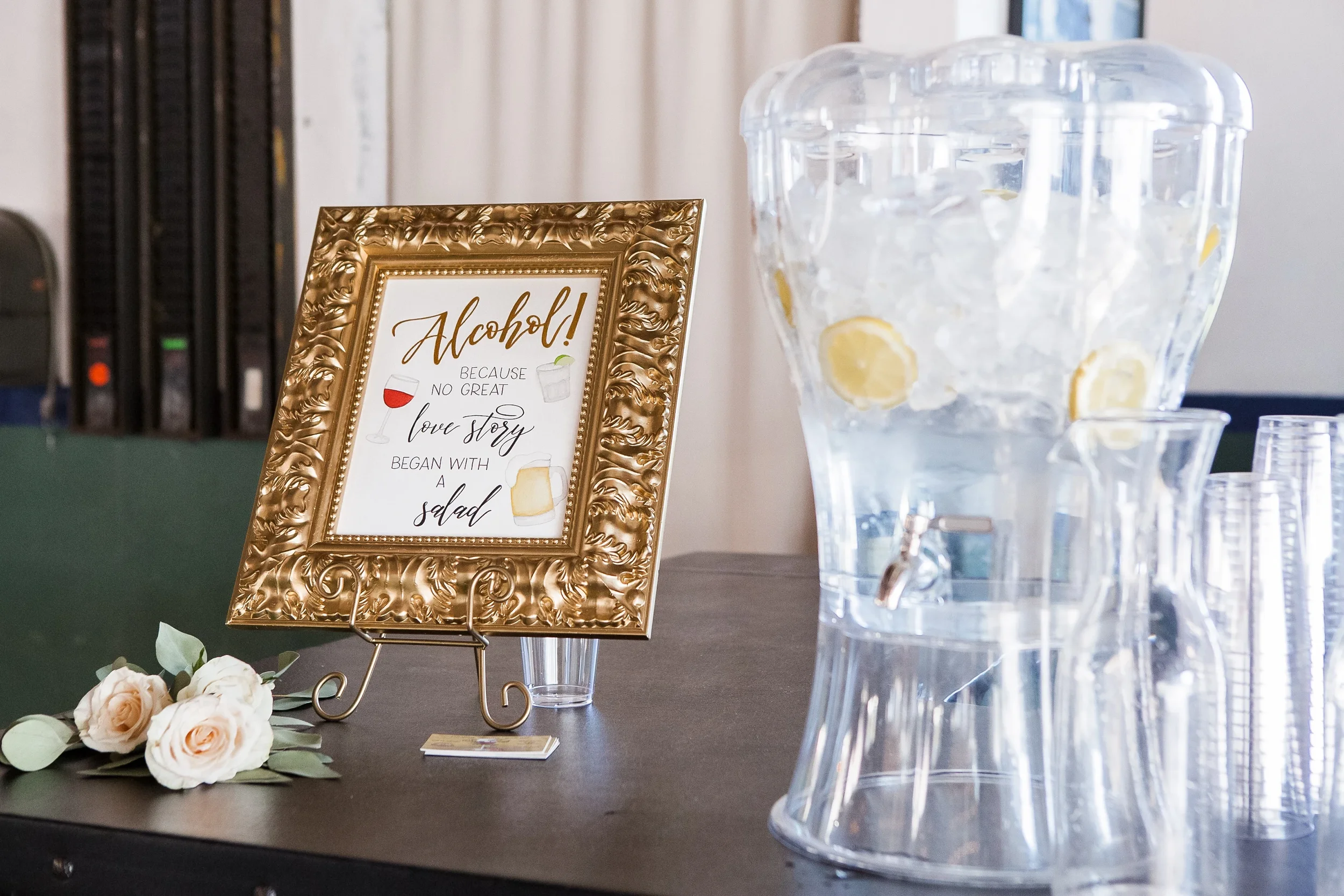 Ornate gold frame drink sign displayed next to a clear glass water dispenser at a wedding reception.