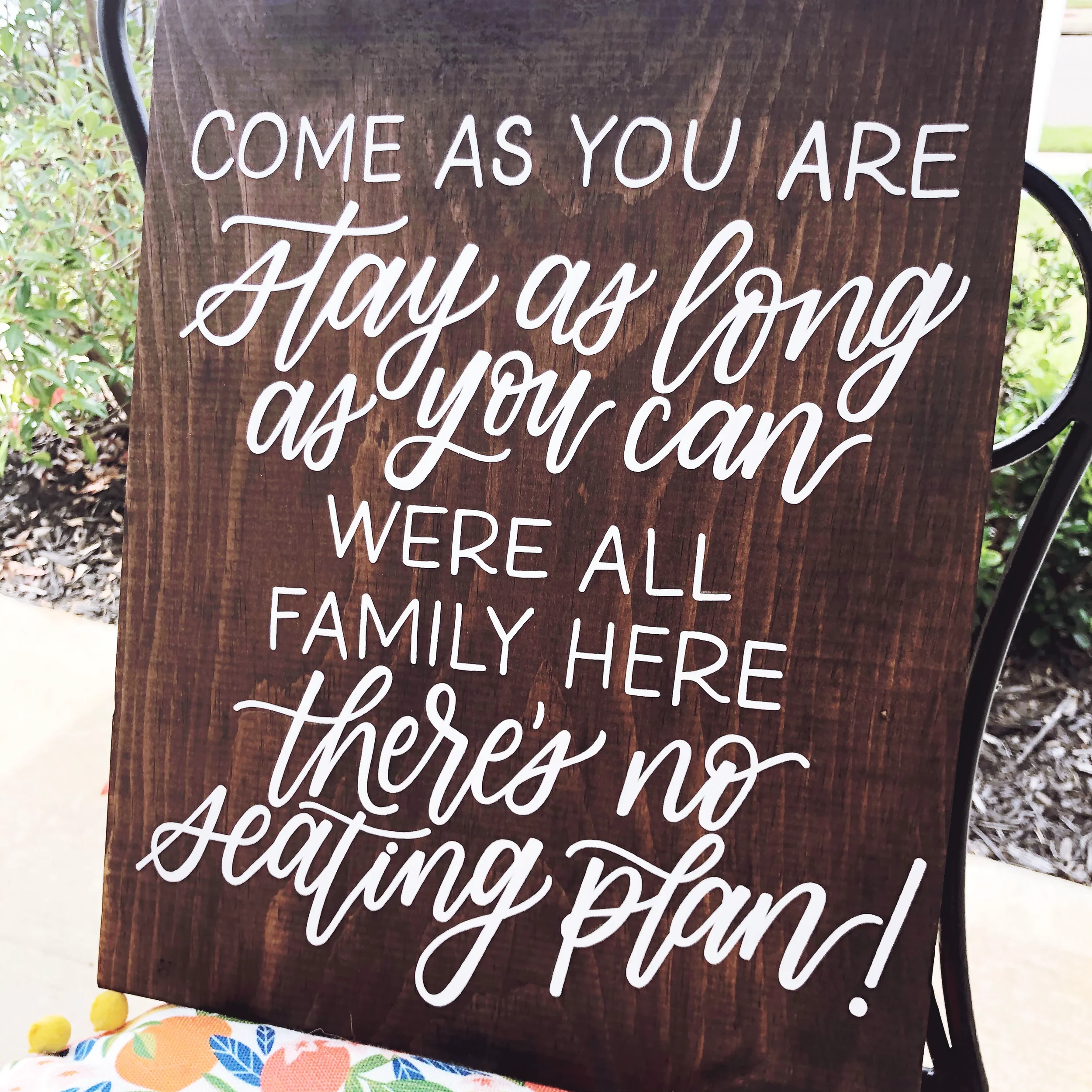 Dark wood sign reading "Come as you are, stay as long as you can, we're all family here, there's no seating plan!" in white cursive script.