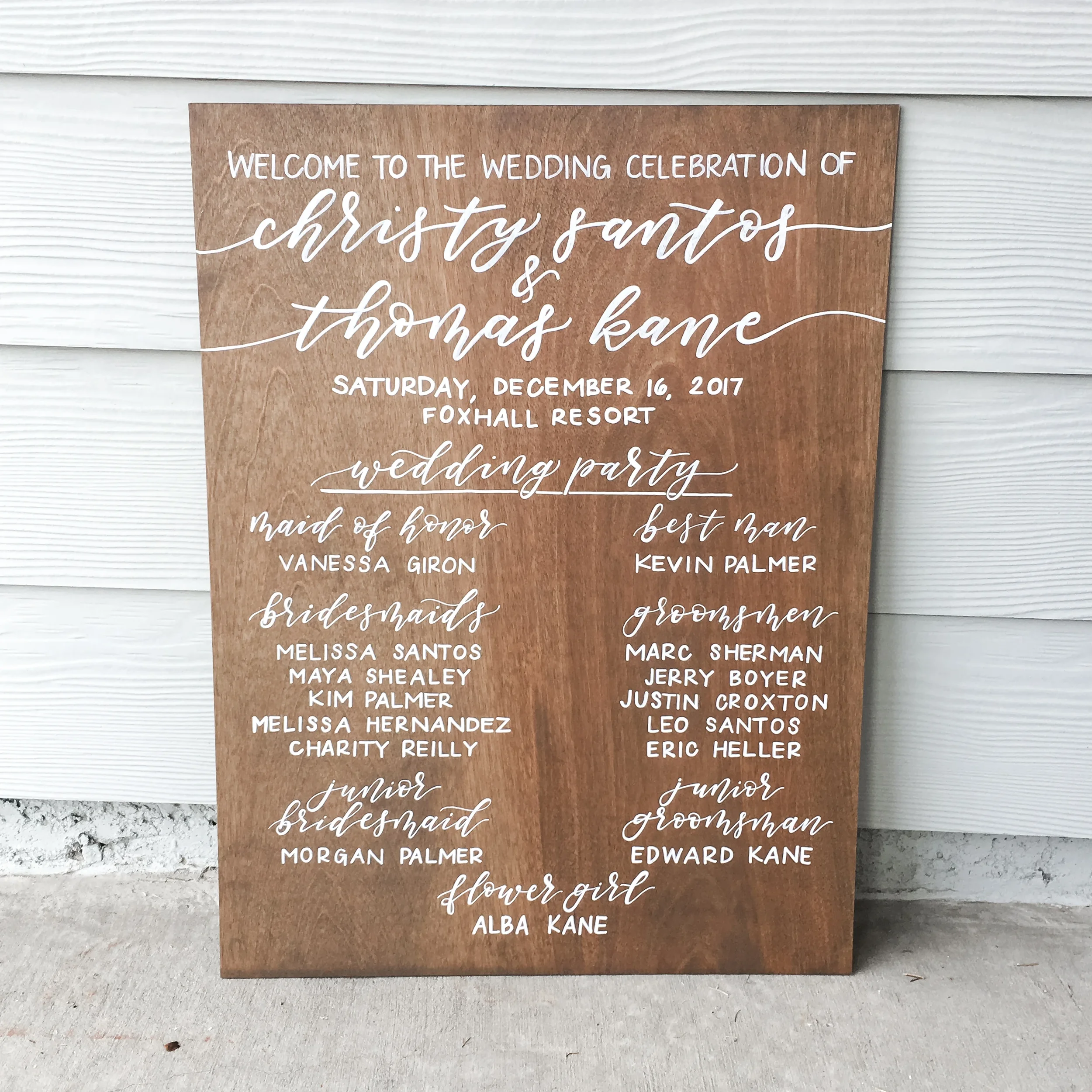 Hand-lettered wooden wedding program sign listing venue and wedding party names.