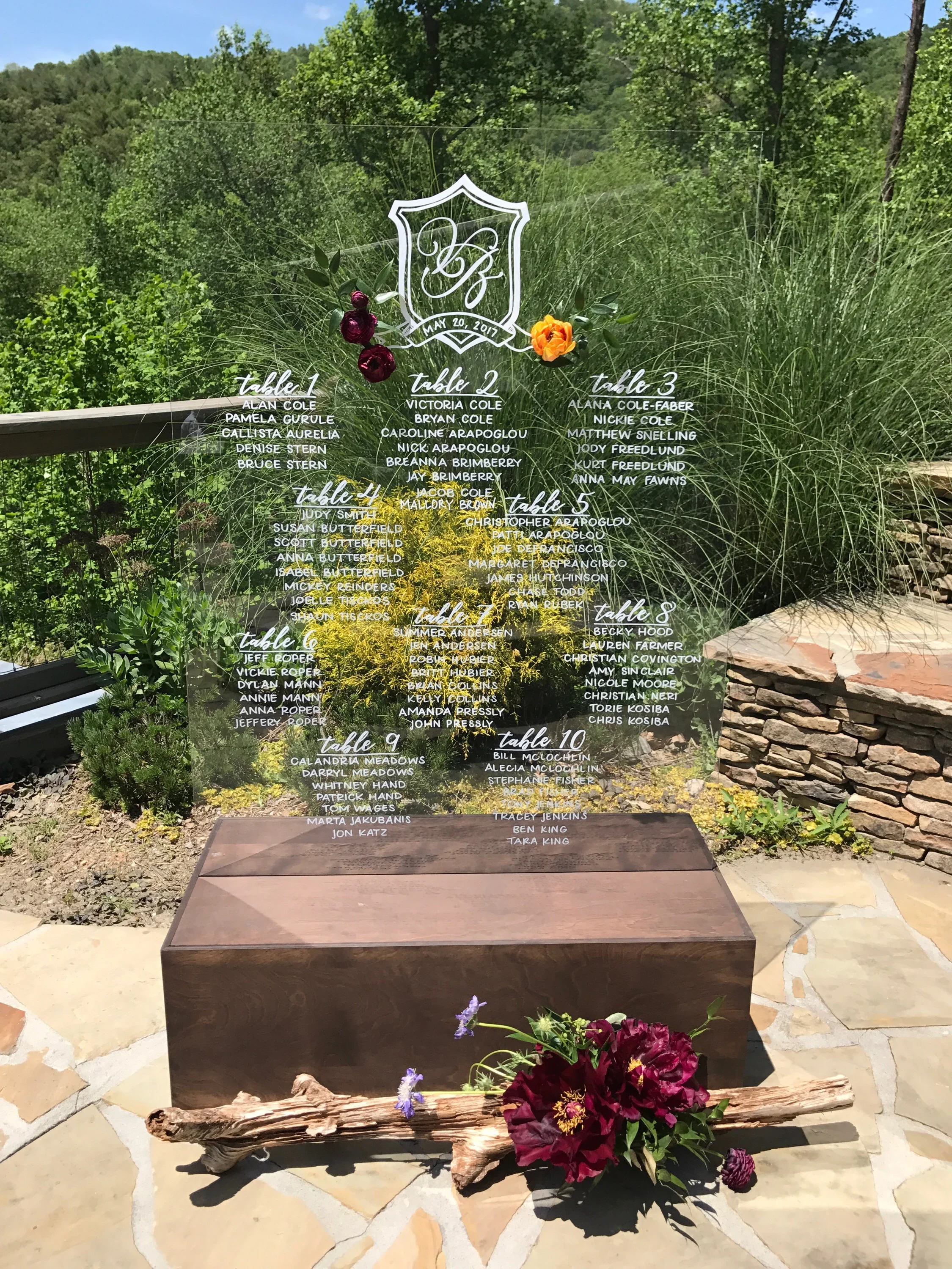 Clear acrylic wedding seating chart board slotted in dark stained wood stand and decorated with deep burgundy florals for an outdoor wedding.