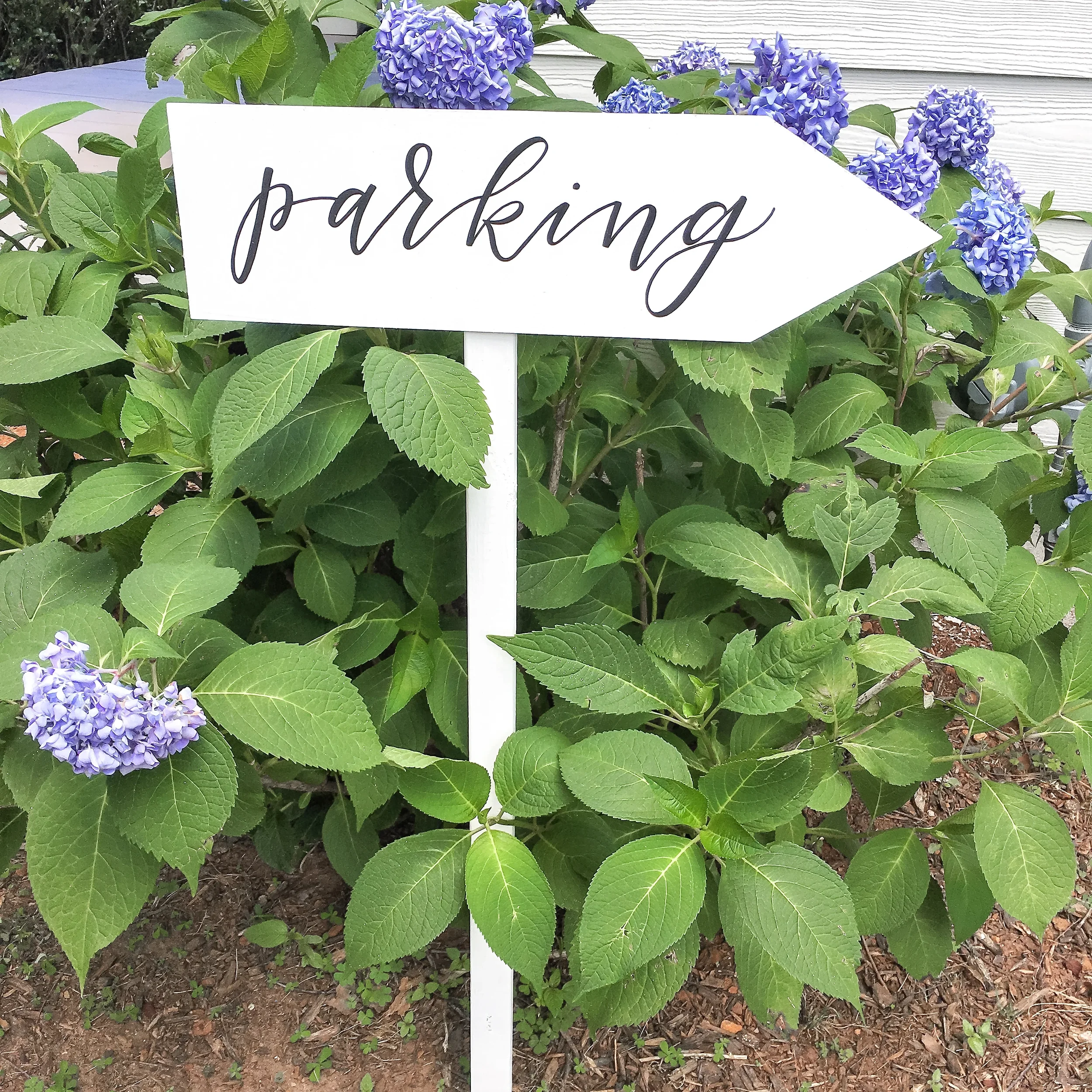 Outdoor white wooden arrow signpost pointing guests toward parking lot at a wedding venue.