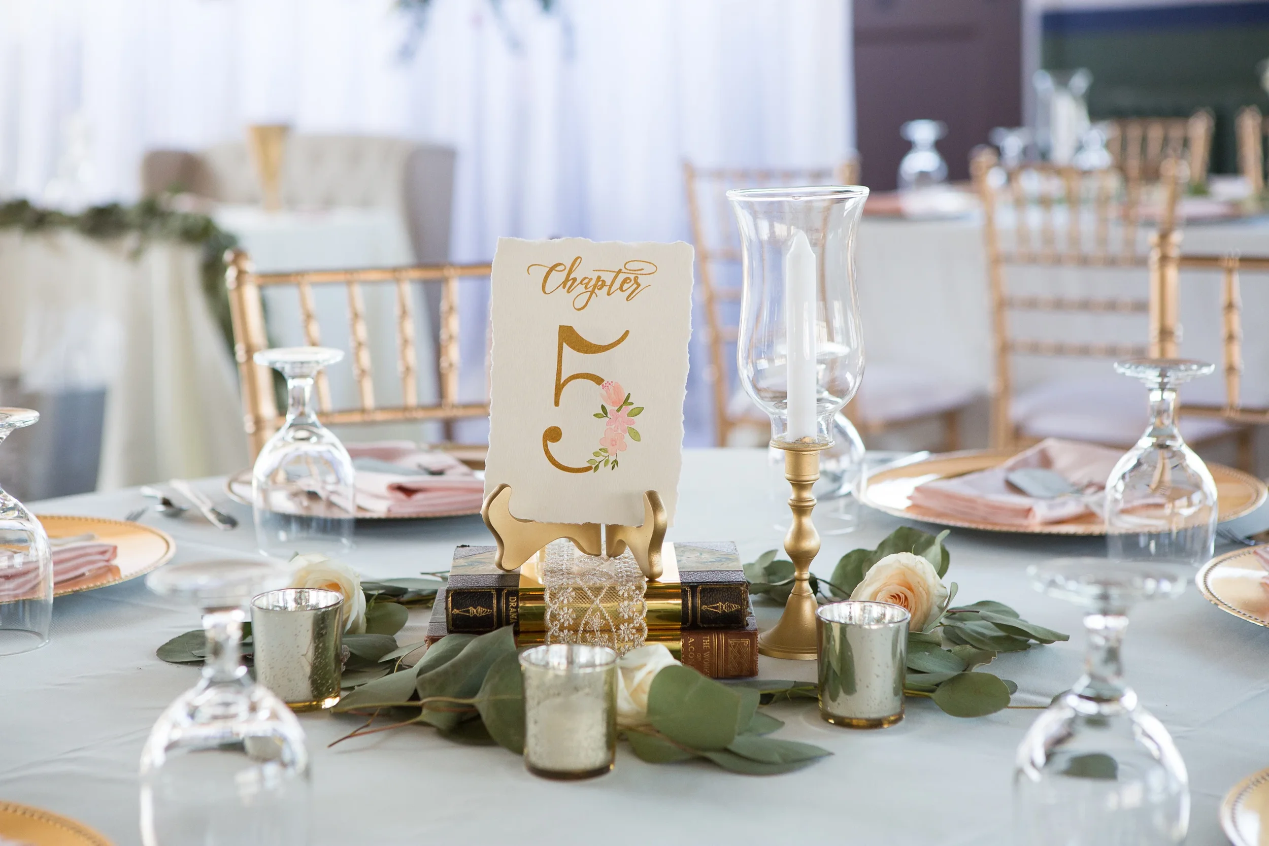 Deckled-edge table number with gold script and painted pink flowers, displayed on vintage books.