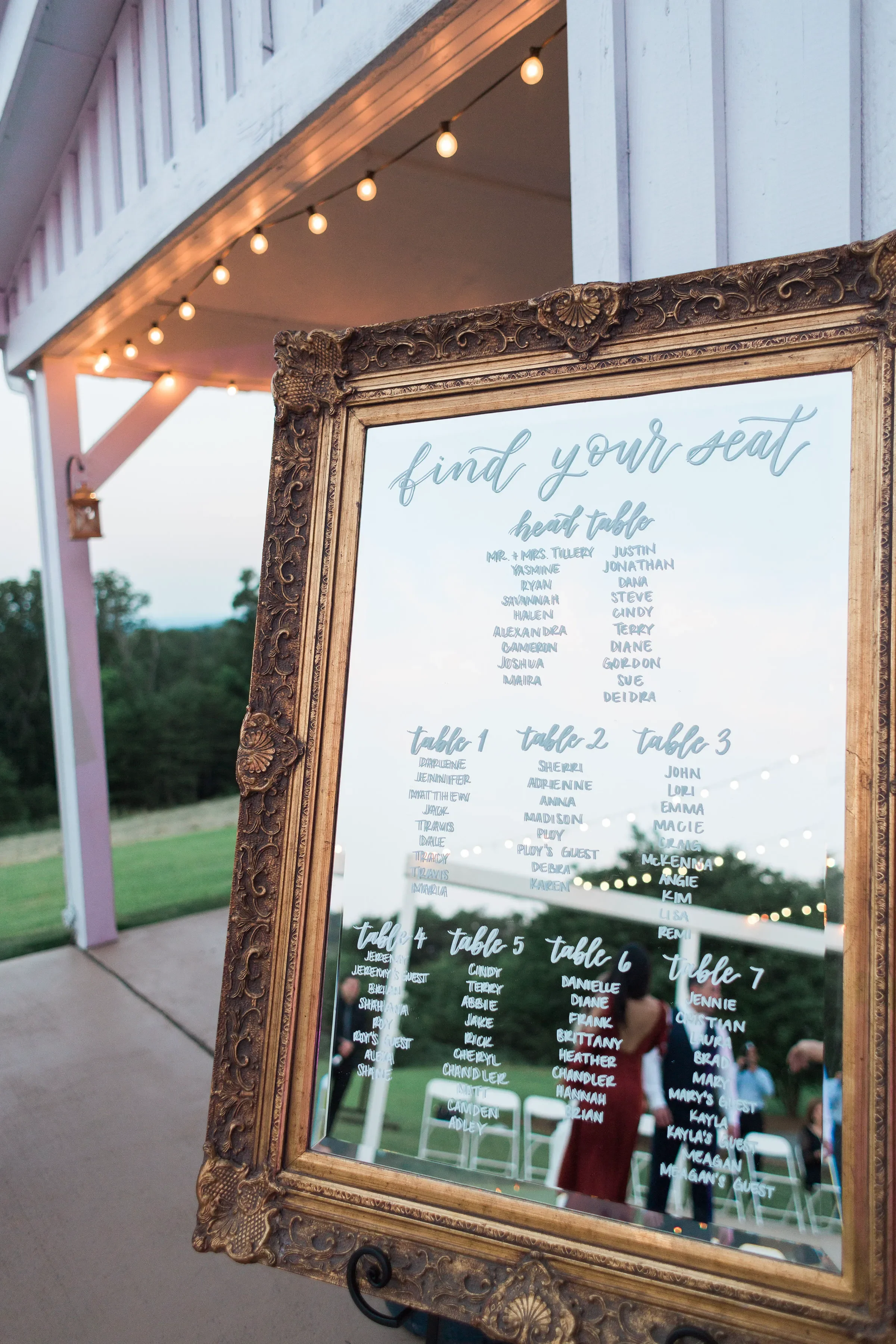 A large vintage gold-framed mirror used as a wedding seating chart with white script calligraphy reading "find your seat" above listed guest tables, displayed outdoors under string lights at an event venue.