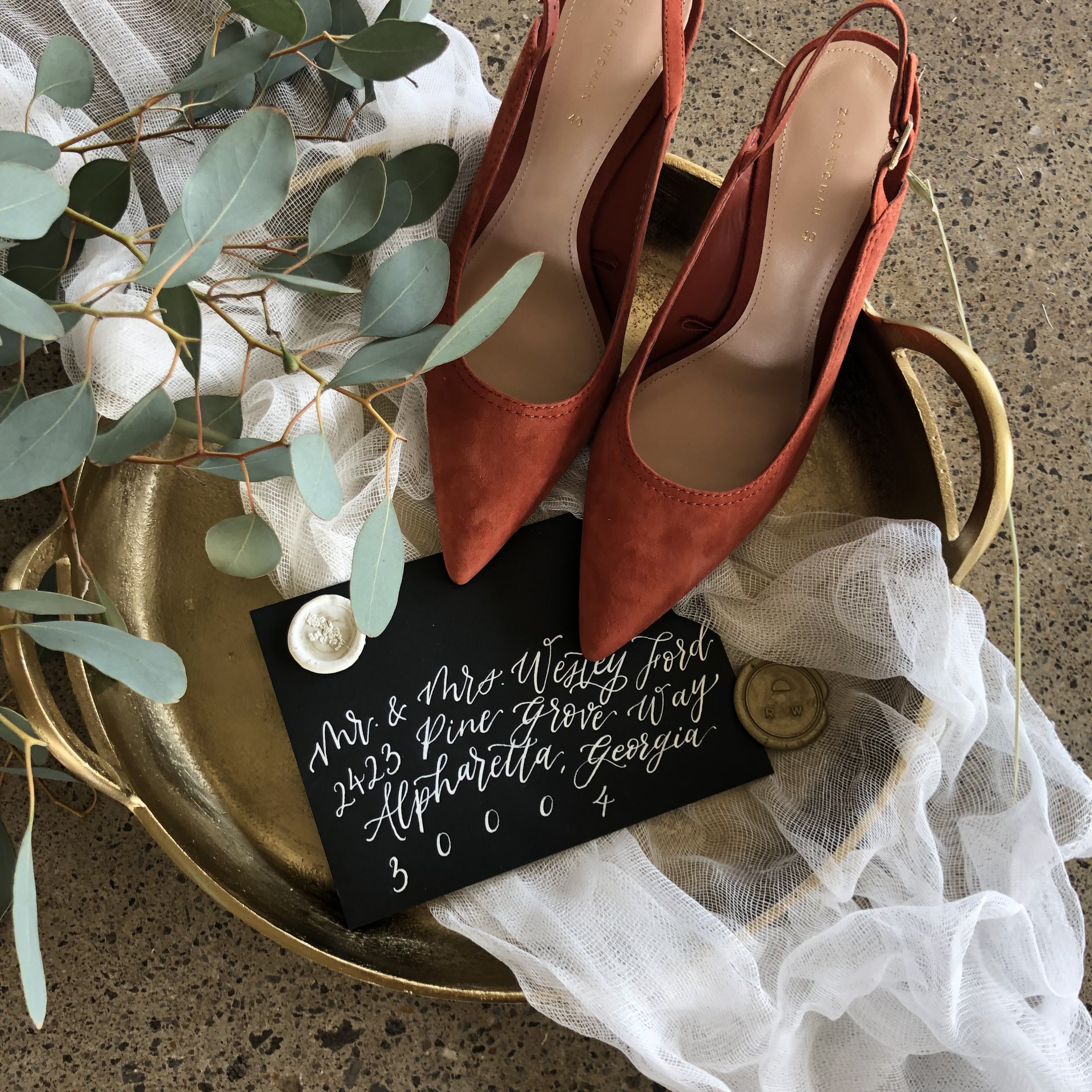 Black wedding envelope with white modern calligraphy address, styled with terracotta bridal shoes, eucalyptus leaves, gold tray, wax seal, and soft white fabric.