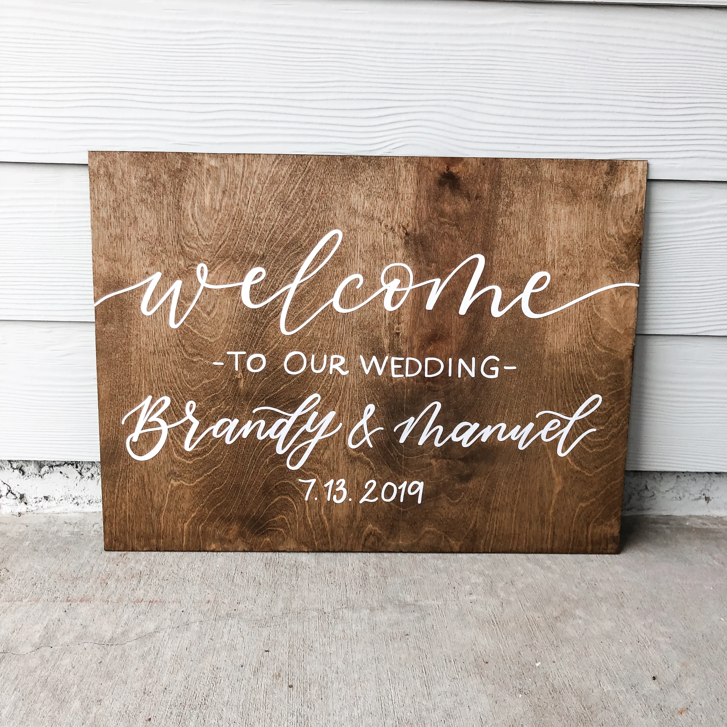 Horizontal stained wood wedding welcome sign hand-lettered in white ink with the couple's names "Brandy & Manuel".