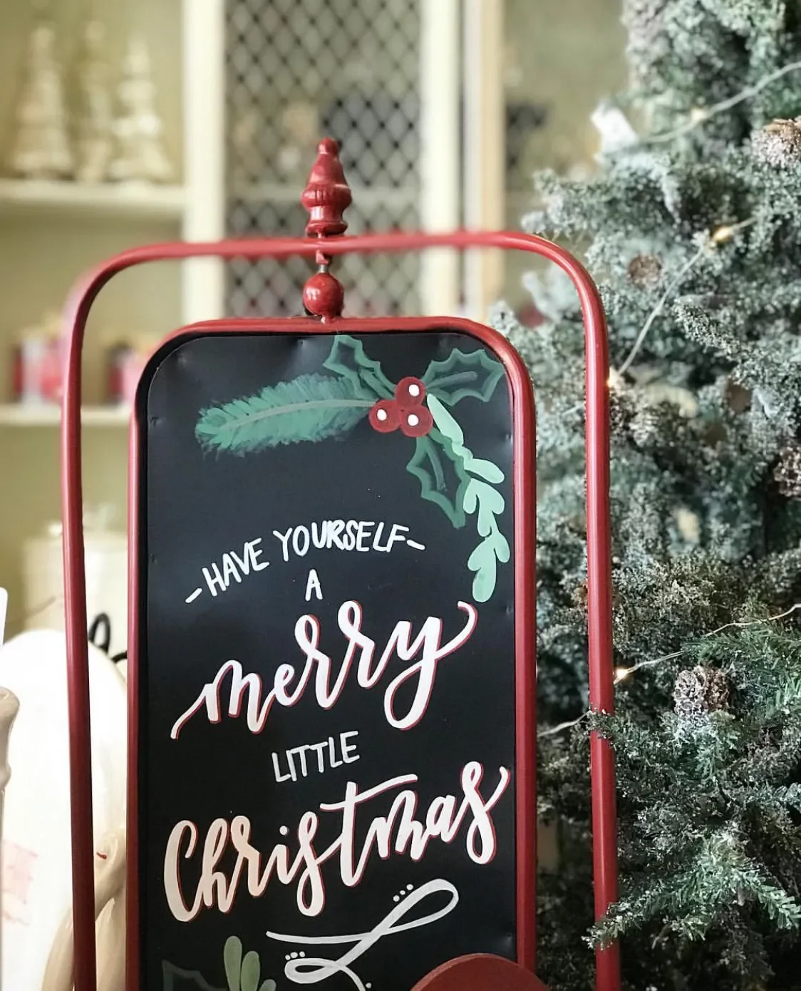 Red framed holiday chalkboard sign reading "Have Yourself a Merry Little Christmas" next to a Christmas tree with hand-painted holly illustrations.
