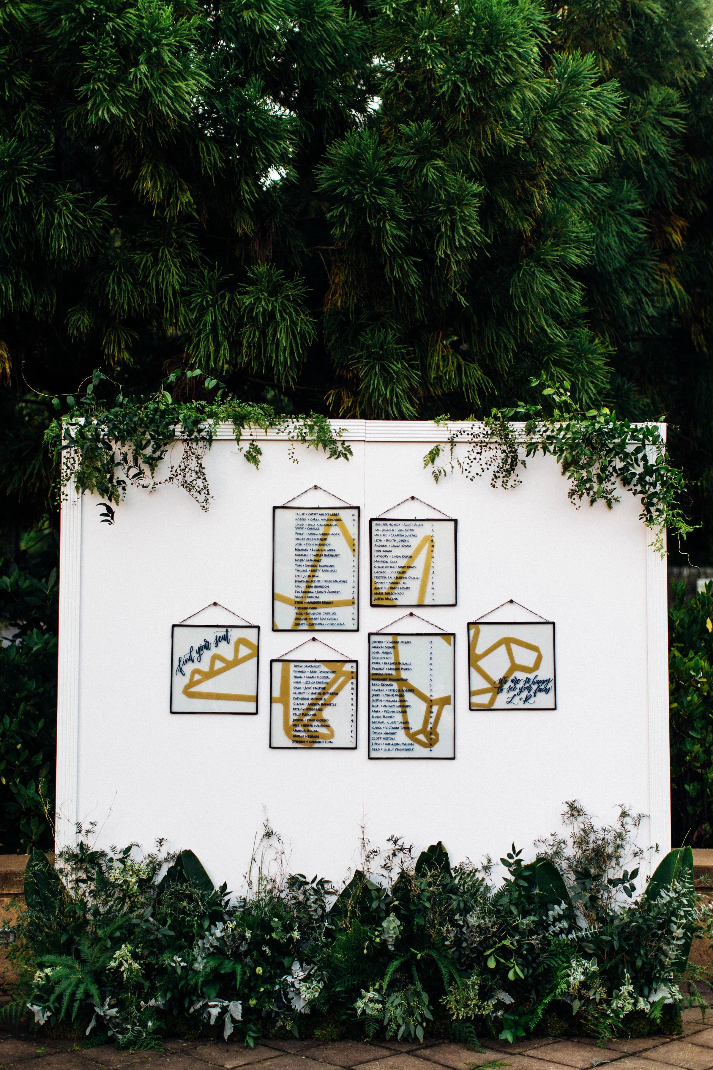 Suspended transparent acrylic and glass frames featuring gold calligraphy seating charts against a large white backdrop.