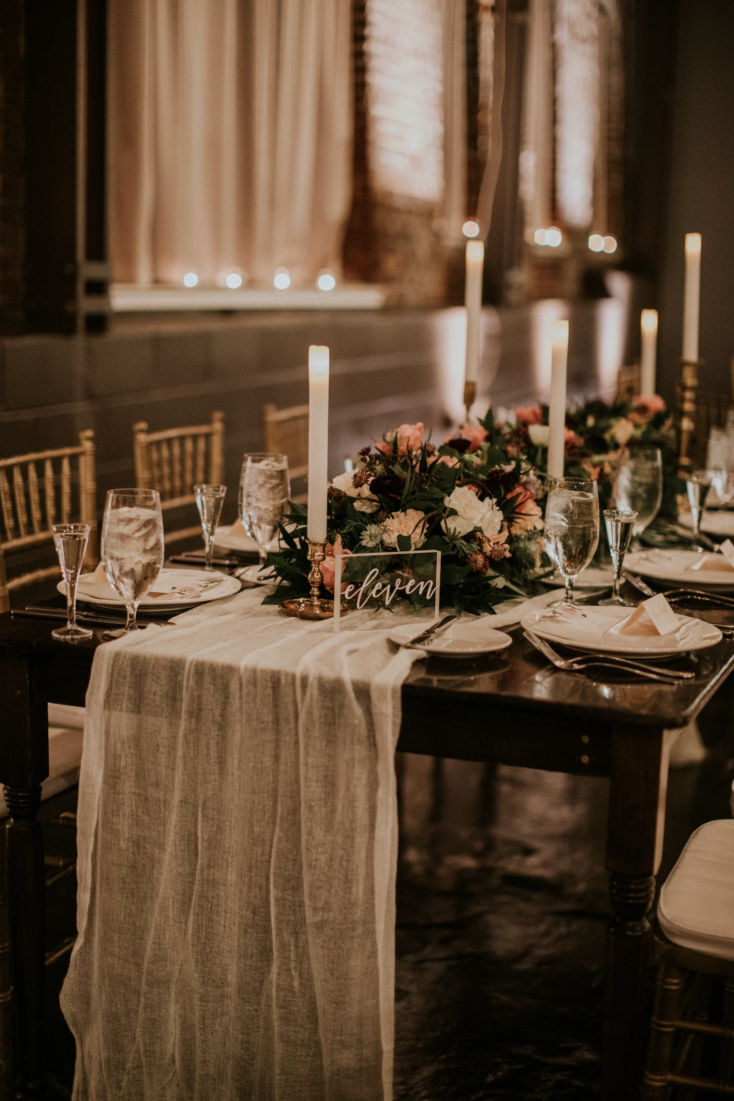 A romantic atmosphere with custom acrylic wedding table numbers featuring elegant white script calligraphy. This clear acrylic table sign reading "eleven" is beautifully styled on a dark wood farm table with an organic white gauze runner, lush garden