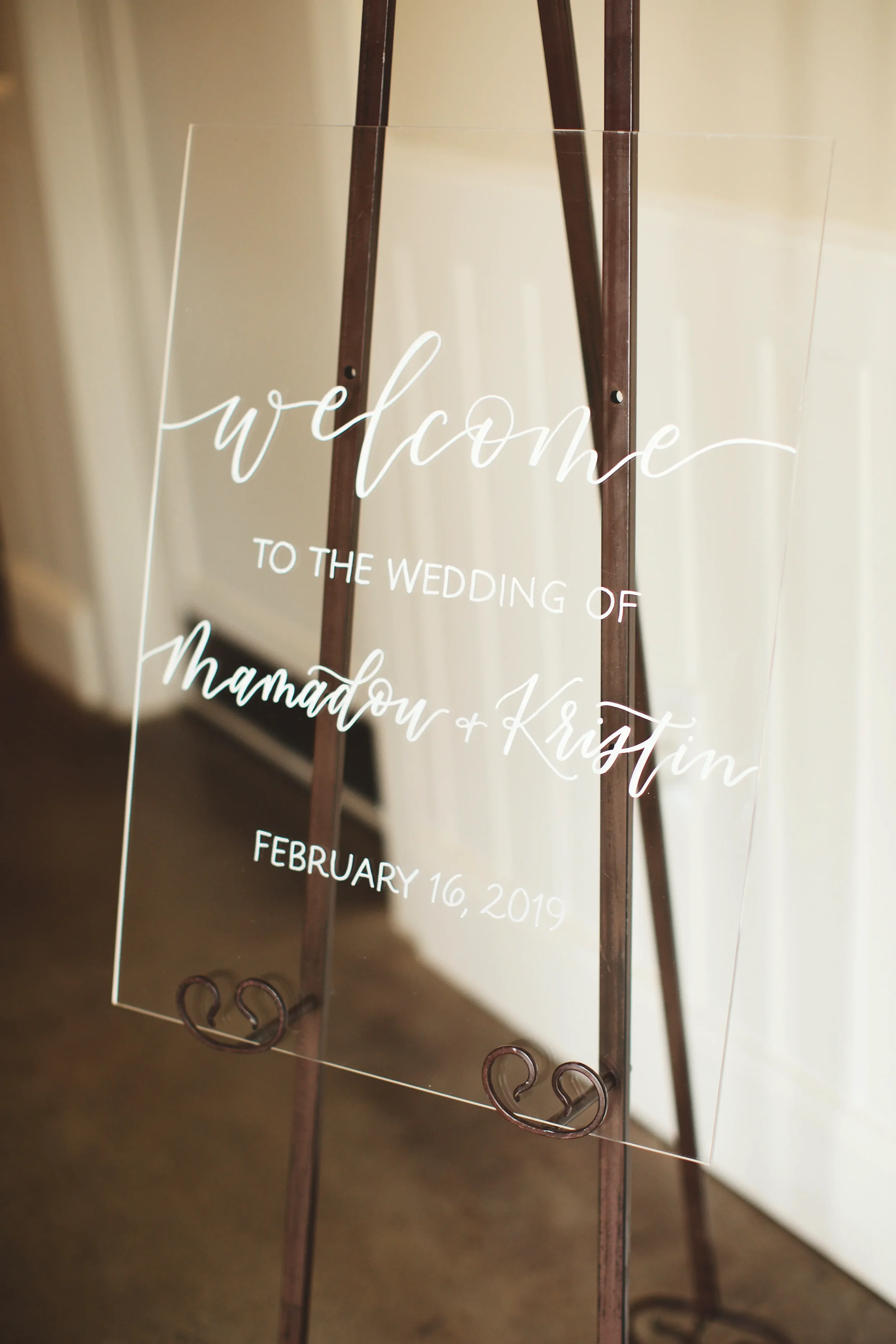 Clear acrylic wedding welcome sign with fine white script calligraphy on a brown metal easel.