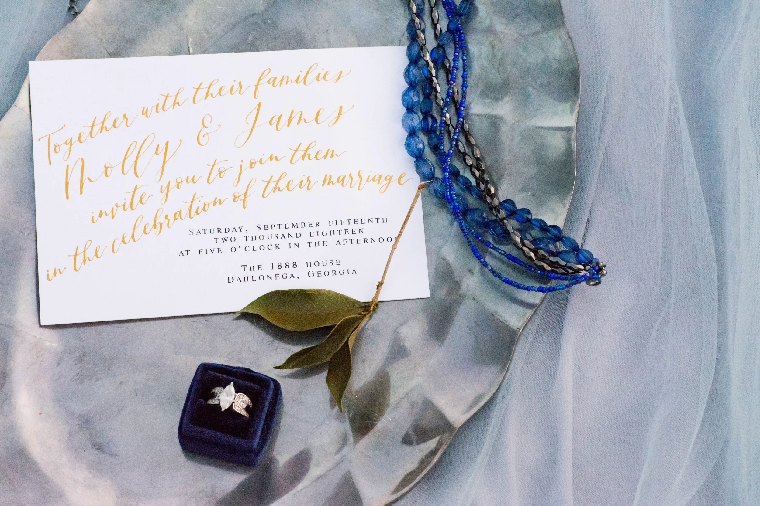 Capture heirloom-quality stationery photos with an artistic flat-lay setup. This design highlights fine gold calligraphy lettering on a classic card, styled over a marble-effect oval platter, a dark velvet ring box, and delicate dusty blue tulle acce