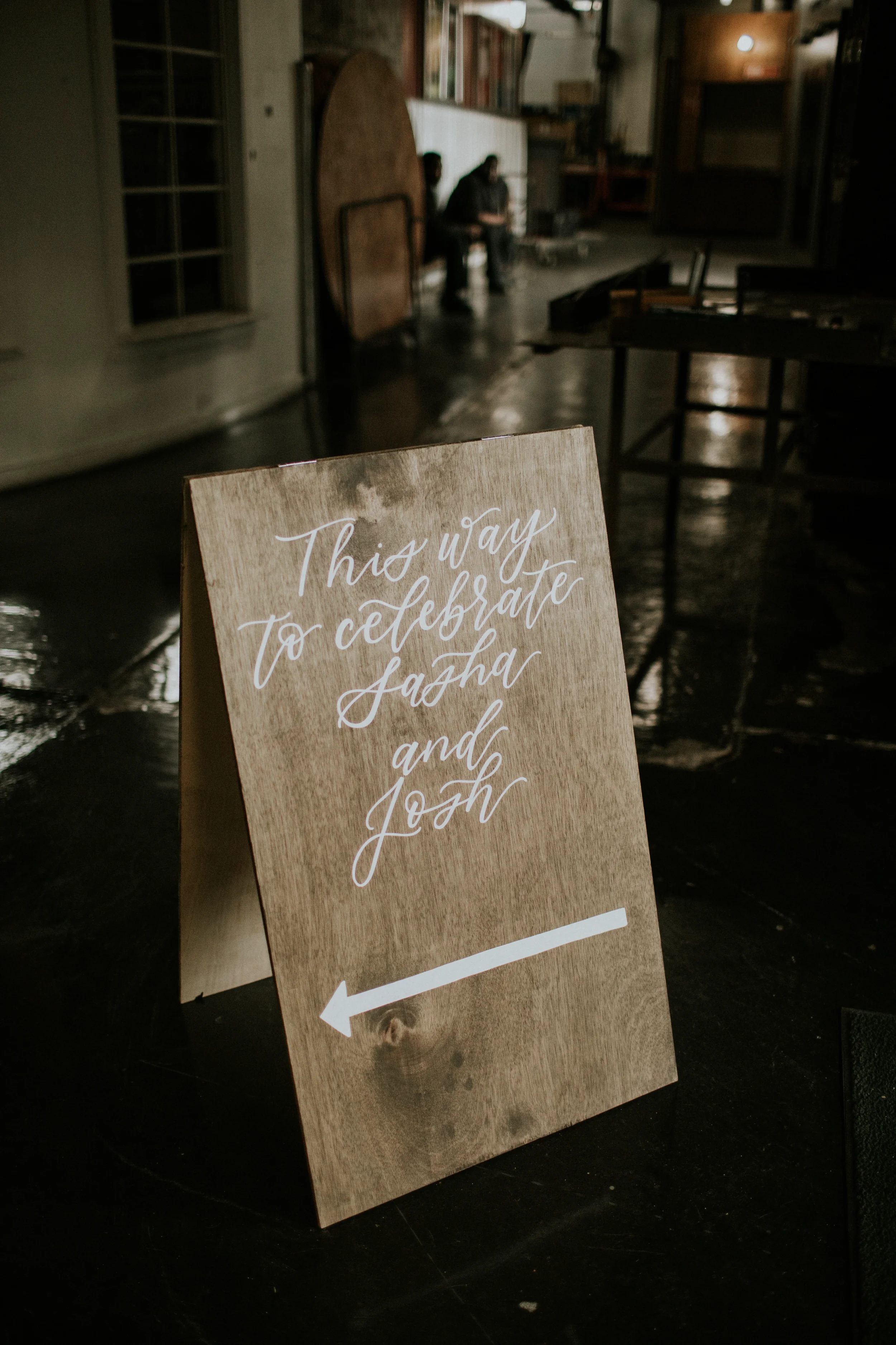 A wooden A-frame sandwich board sign with white script calligraphy reading "This way to celebrate Sasha and Josh" above a left-pointing arrow inside an industrial indoor wedding venue.