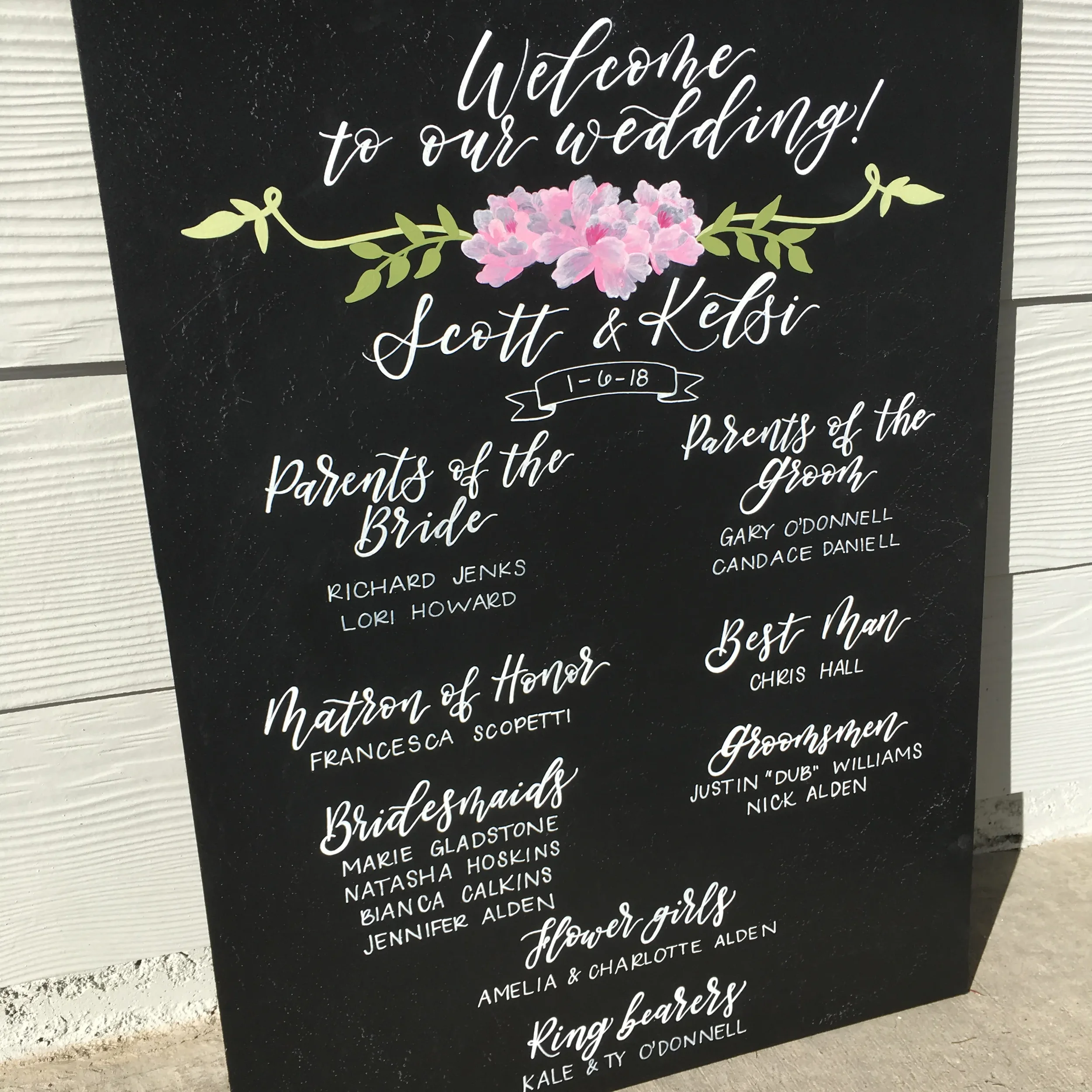 Large chalkboard wedding welcome sign detailing the bridal party names with pink floral chalk art.