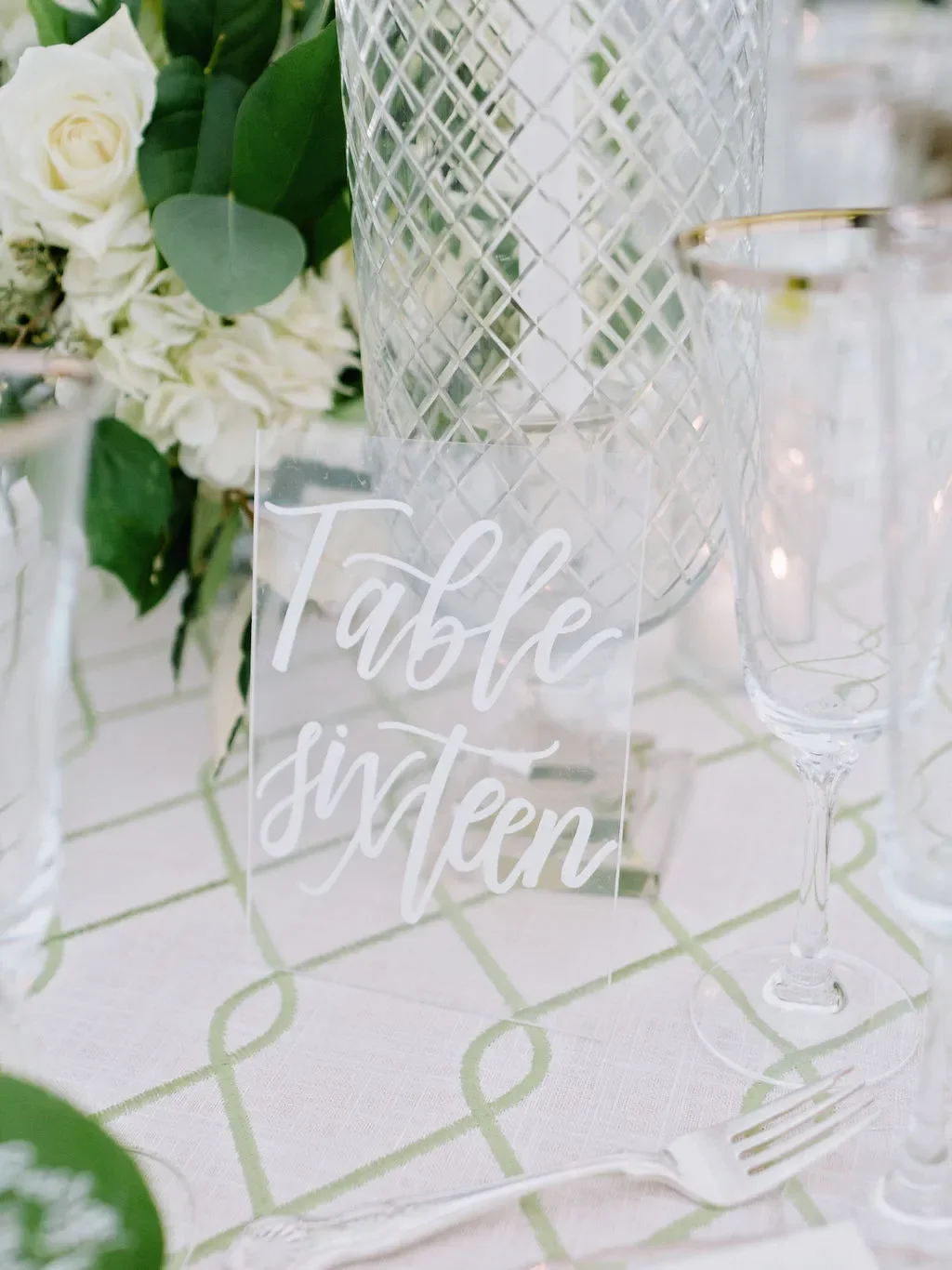 Elegant wedding reception table setting with acrylic table number sign reading "Table Sixteen," white hydrangea and garden rose centerpiece, silver-rimmed champagne flutes, and white pillar candle in glass hurricane holder.