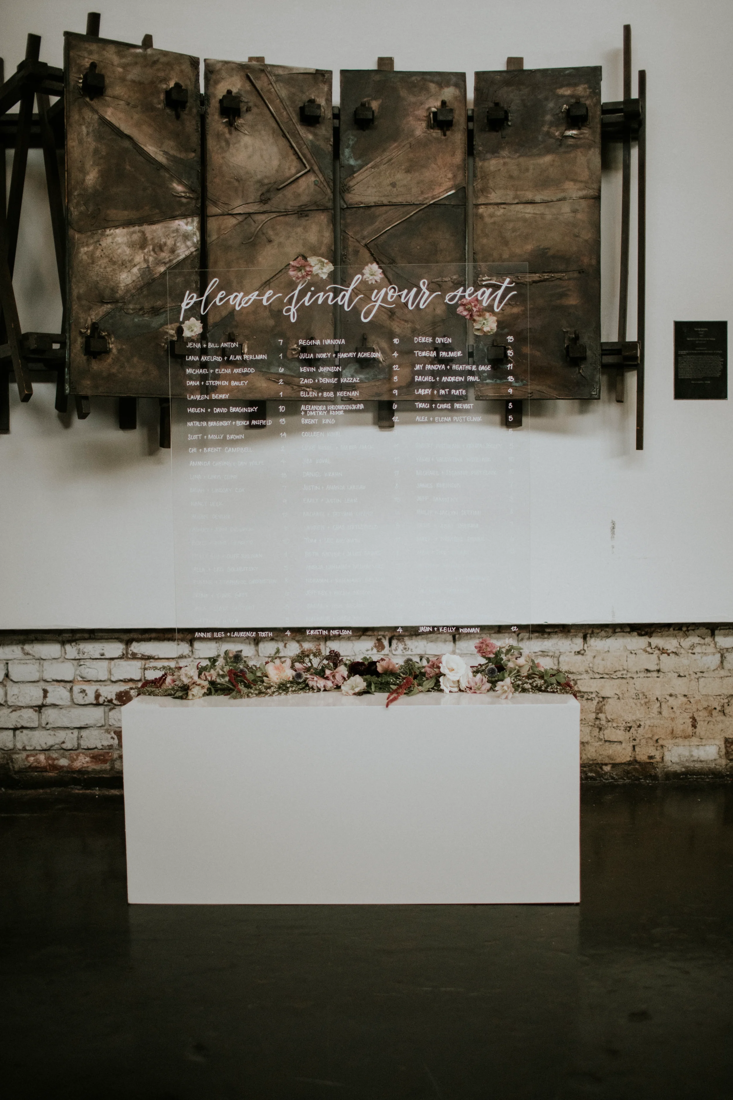 Large clear acrylic wedding seating chart with white script calligraphy reading "please find your seat" resting on a white rectangular block with a floral runner in front of an exposed brick wall.