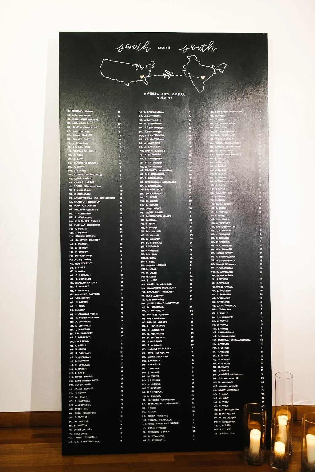 Large black vertical wedding seating chart for love abroad against a white wall with candles.