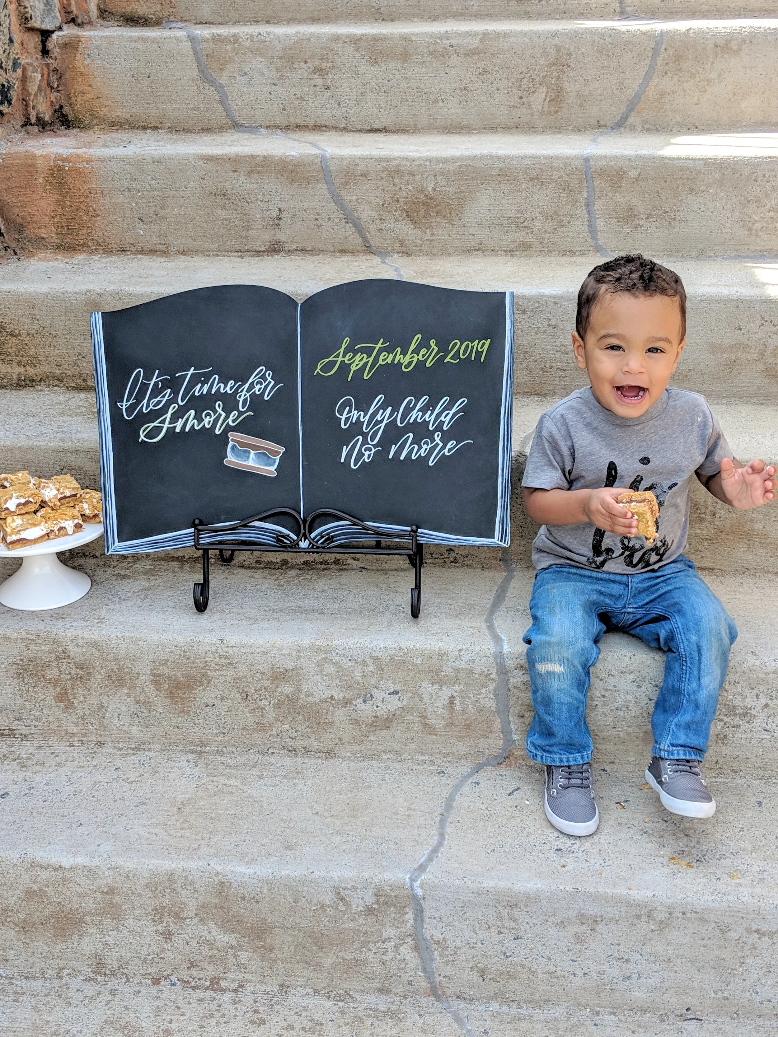 Toddler sitting on concrete steps next to an open-book shaped chalkboard pregnancy announcement sign.