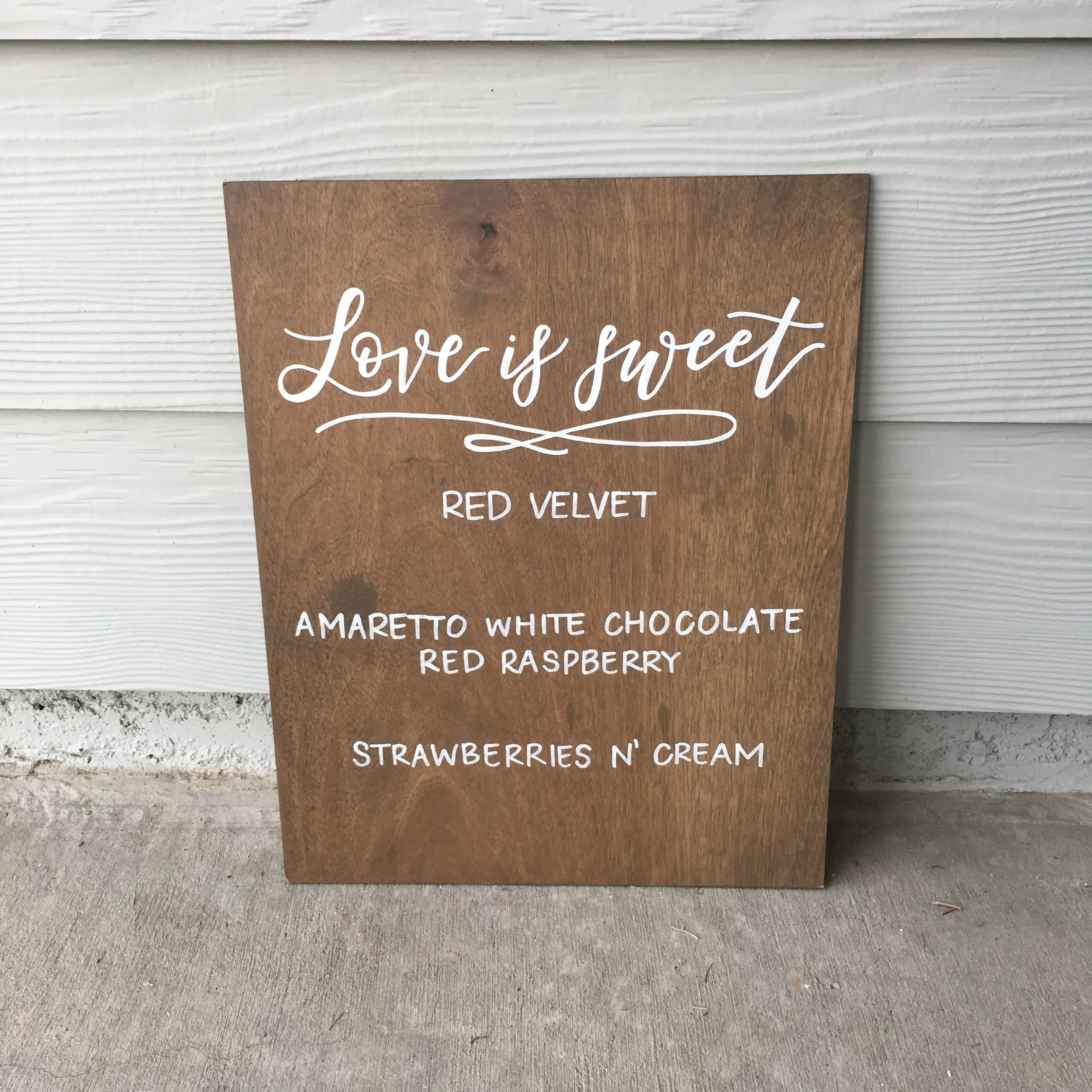 Stained wood dessert sign reading "Love is sweet" with listed cake flavors Red Velvet, Amaretto White Chocolate, and Strawberries n' Cream.