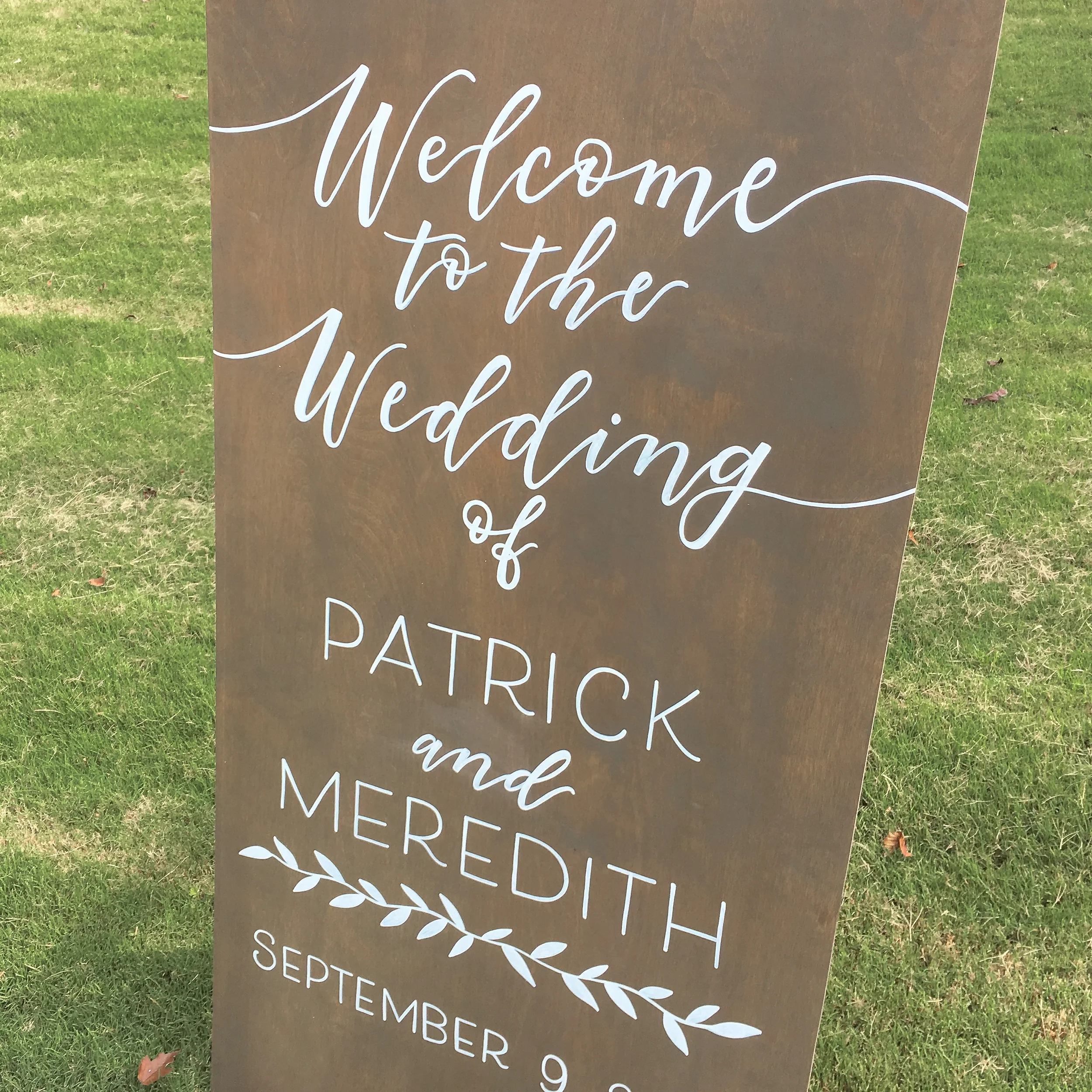 Tall weathered grey-stained wood welcome sign reading "Welcome to the Wedding of Patrick and Meredith" with botanical branch accents.