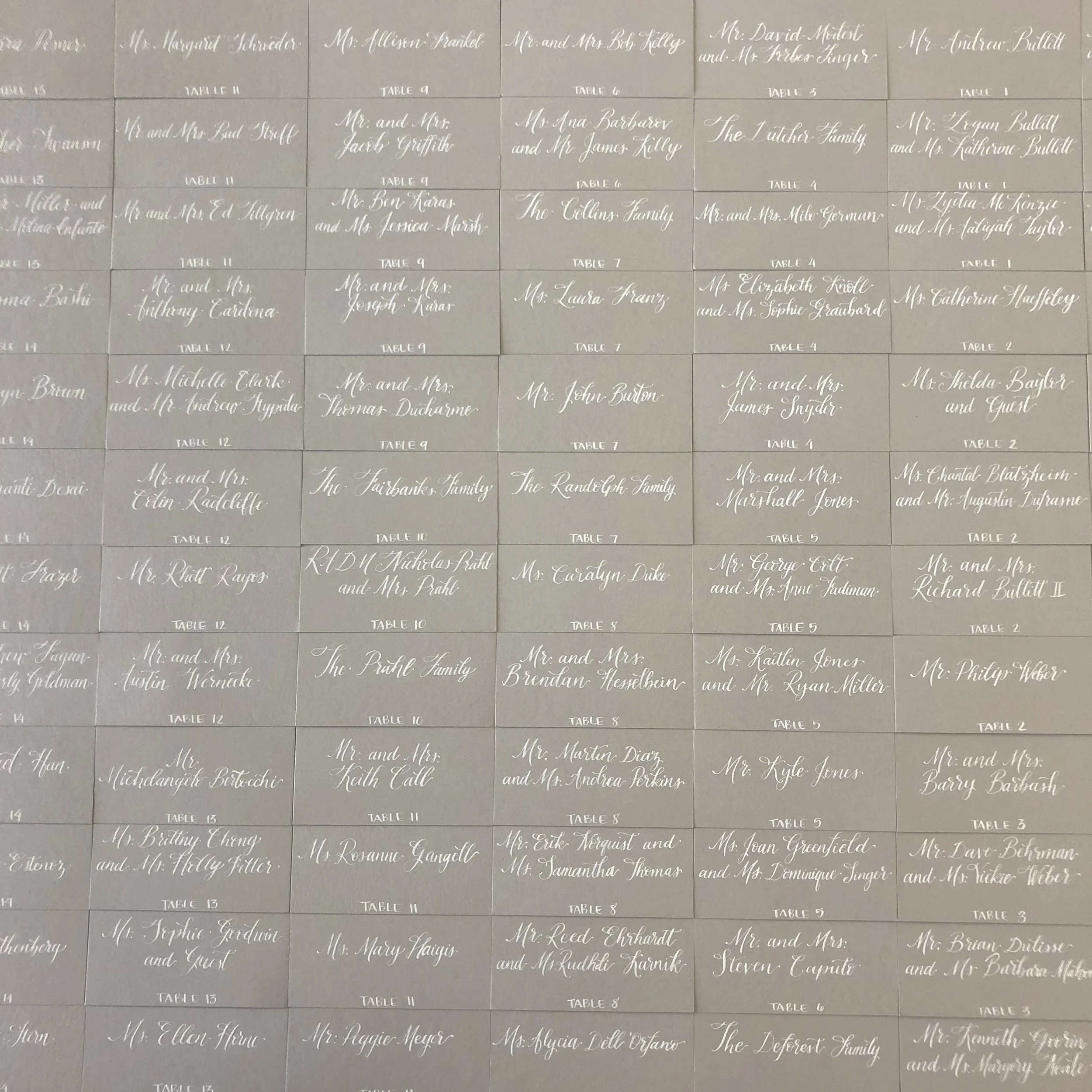 Grid of slate grey escort cards neatly arranged with white calligraphy hand lettering detailing guest names and table numbers.