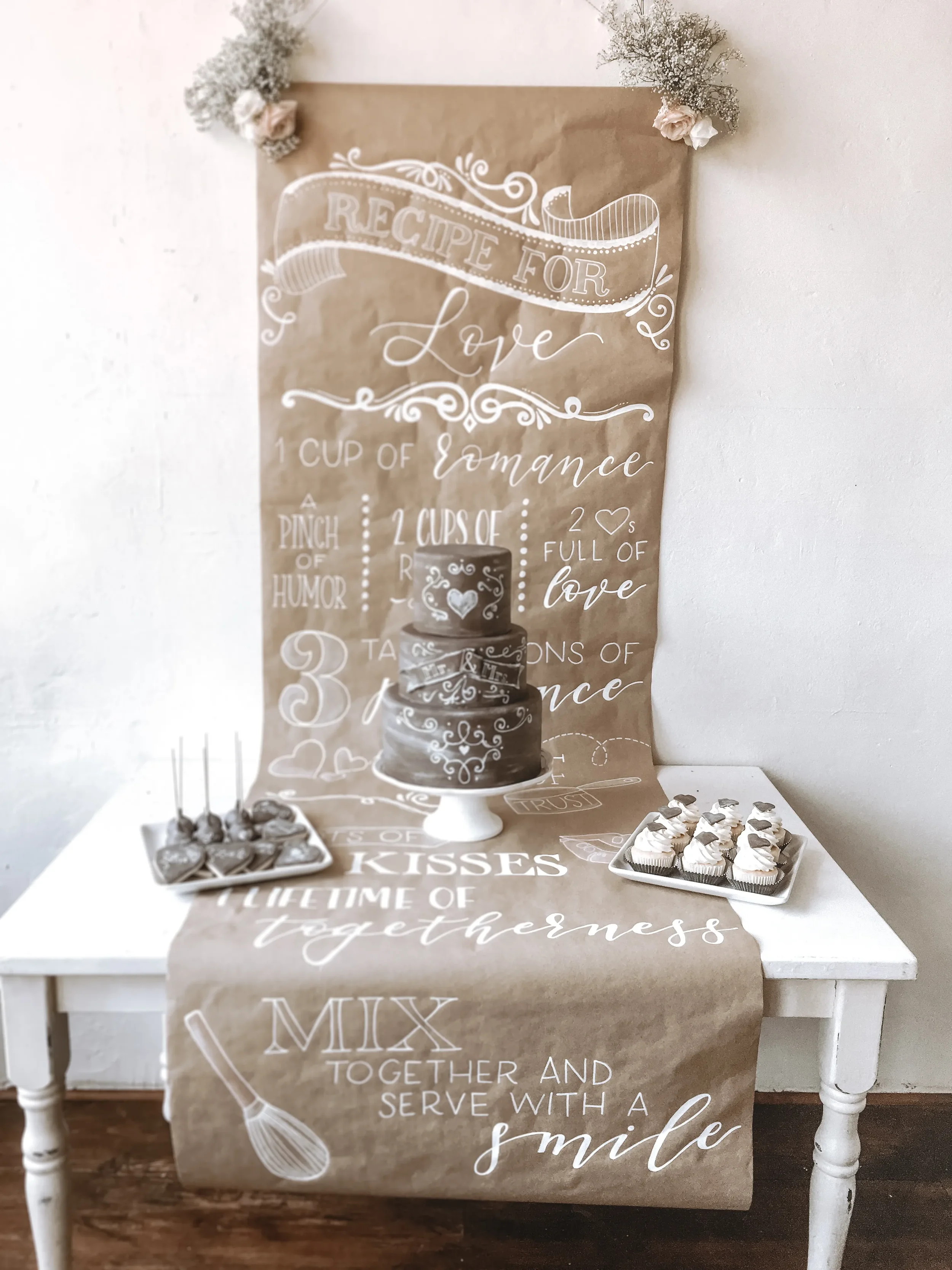 Hand-lettered brown kraft paper table runner reading "Recipe for Love" draped over a wedding dessert table.