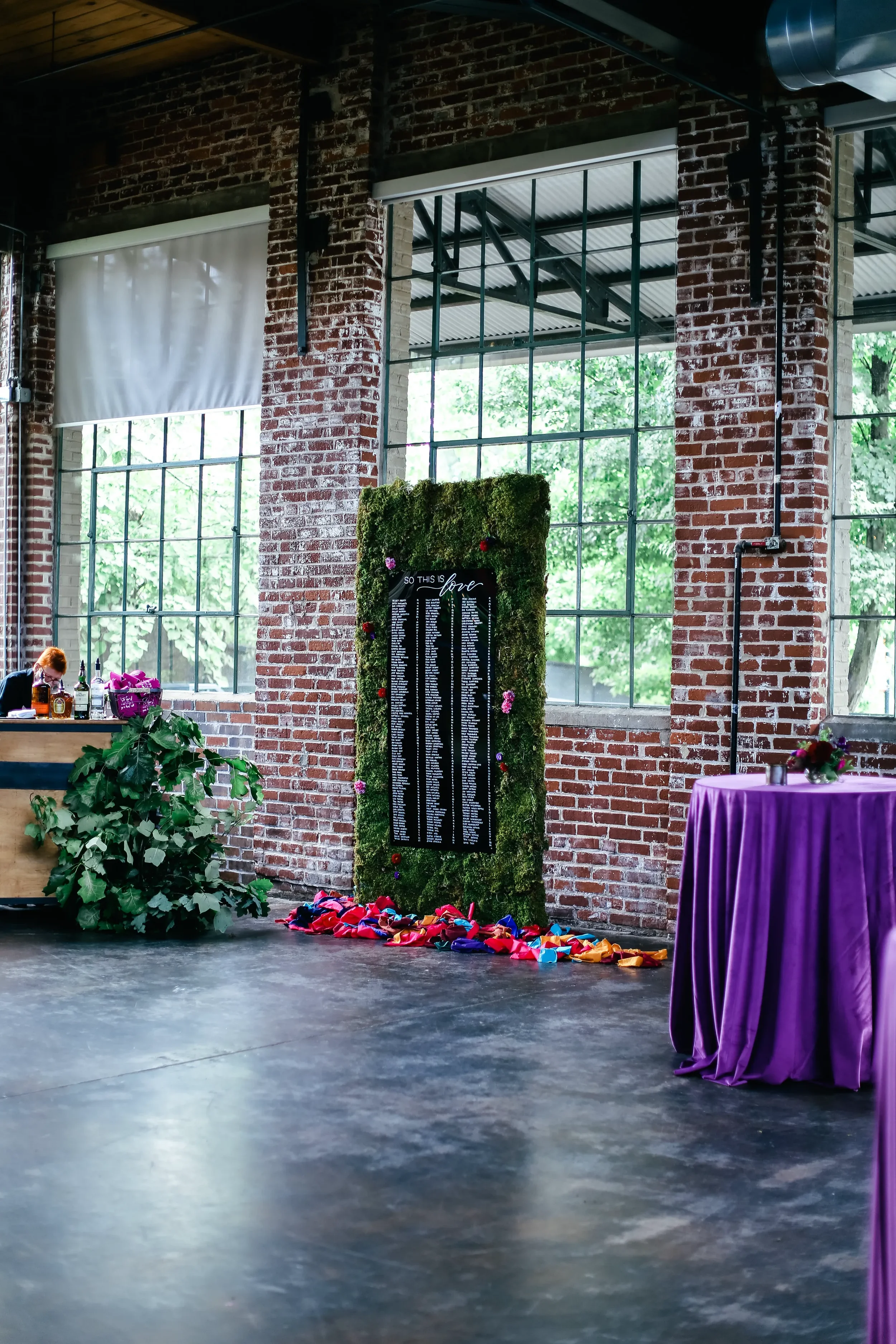 Create an jaw-dropping centerpiece in an industrial brick venue with a statement living moss wall seating chart. The high-contrast black acrylic directory features precise white text and romantic calligraphy accents, framed perfectly by organic moss,