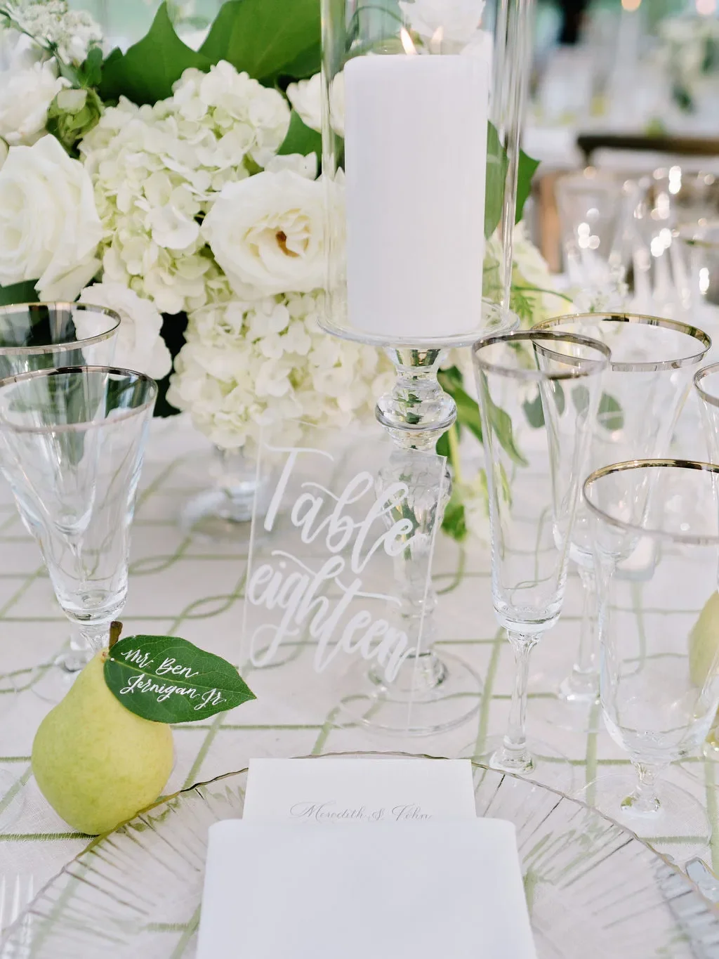 Clear acrylic table number sign with white calligraphy, styled with a pear escort card, white floral centerpiece, and elegant wedding reception table decor.
