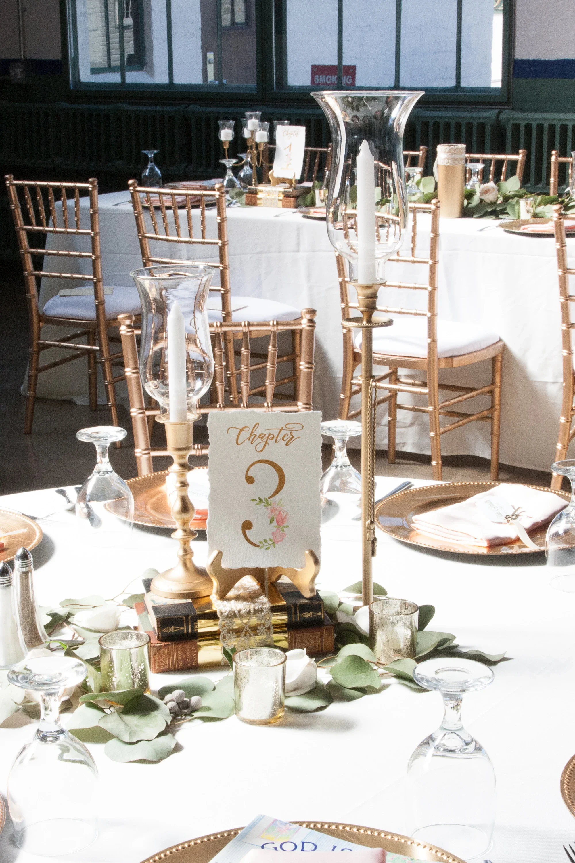 Romantic floral-illustrated wedding table sign placed on a gold easel over stacked antique books.