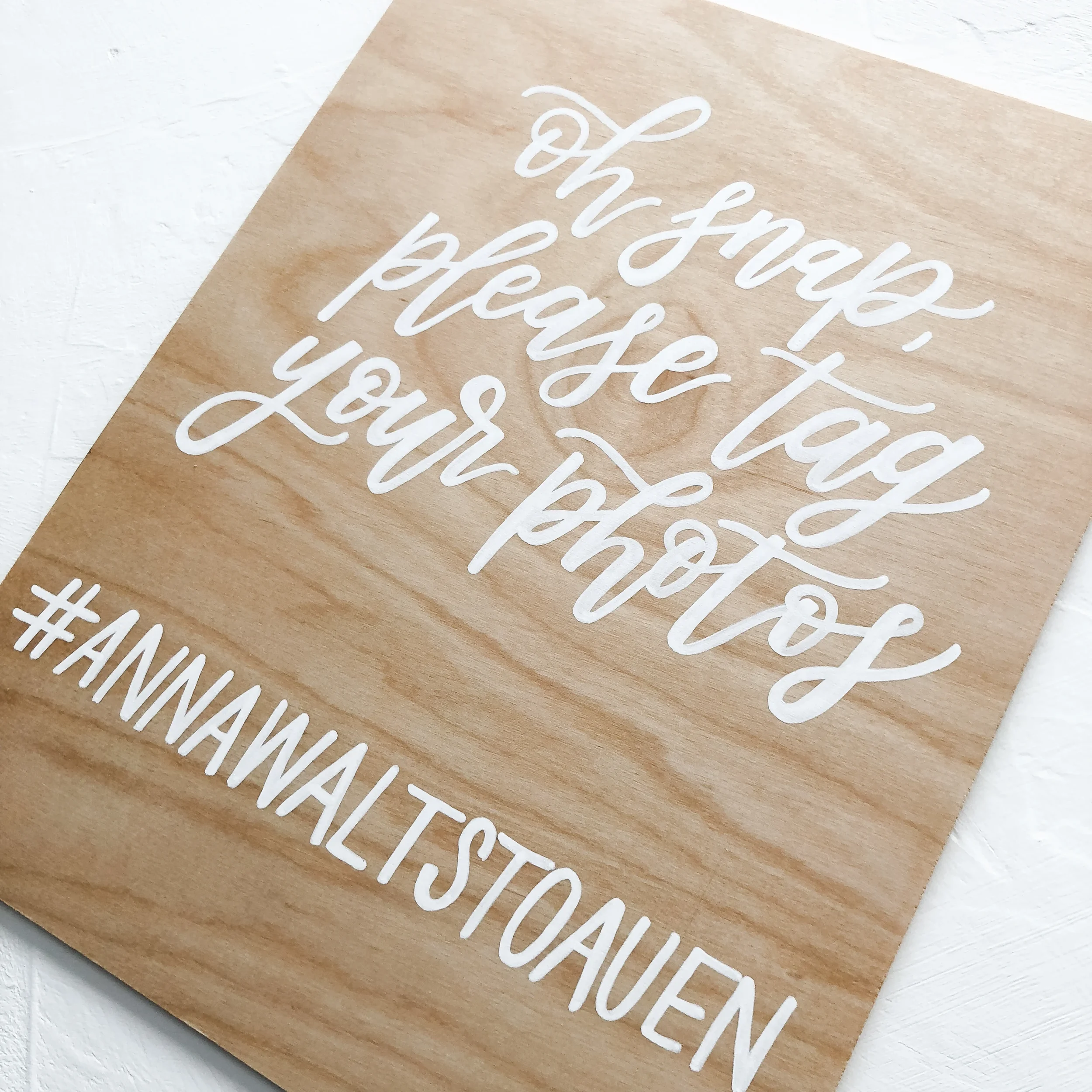Natural light wood sign on a mini easel featuring white calligraphy that reads "Oh snap, please tag your photos" with a custom wedding hashtag.
