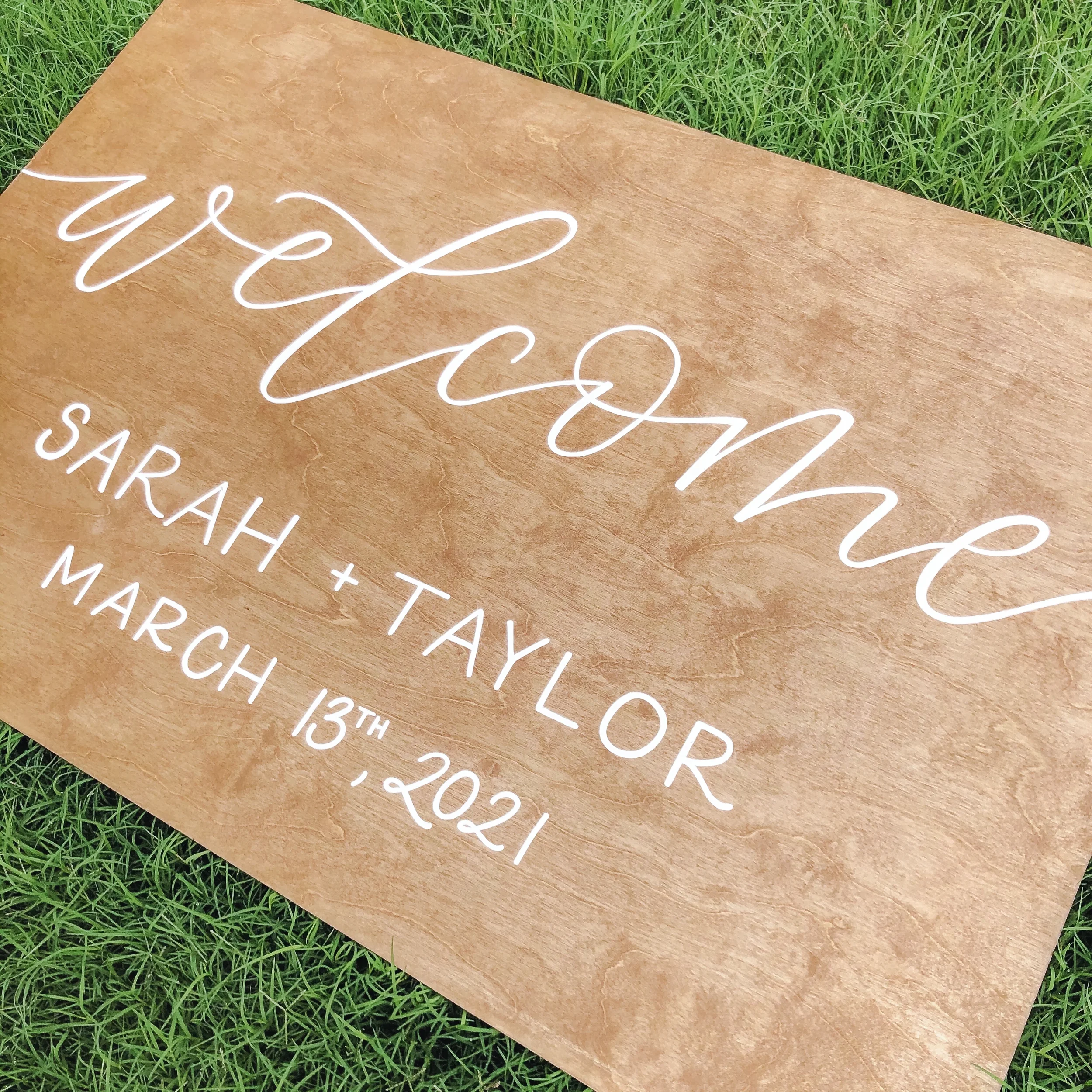 Horizontal stained wood wedding welcome sign hand-lettered in white ink with the couple's names and wedding date.
