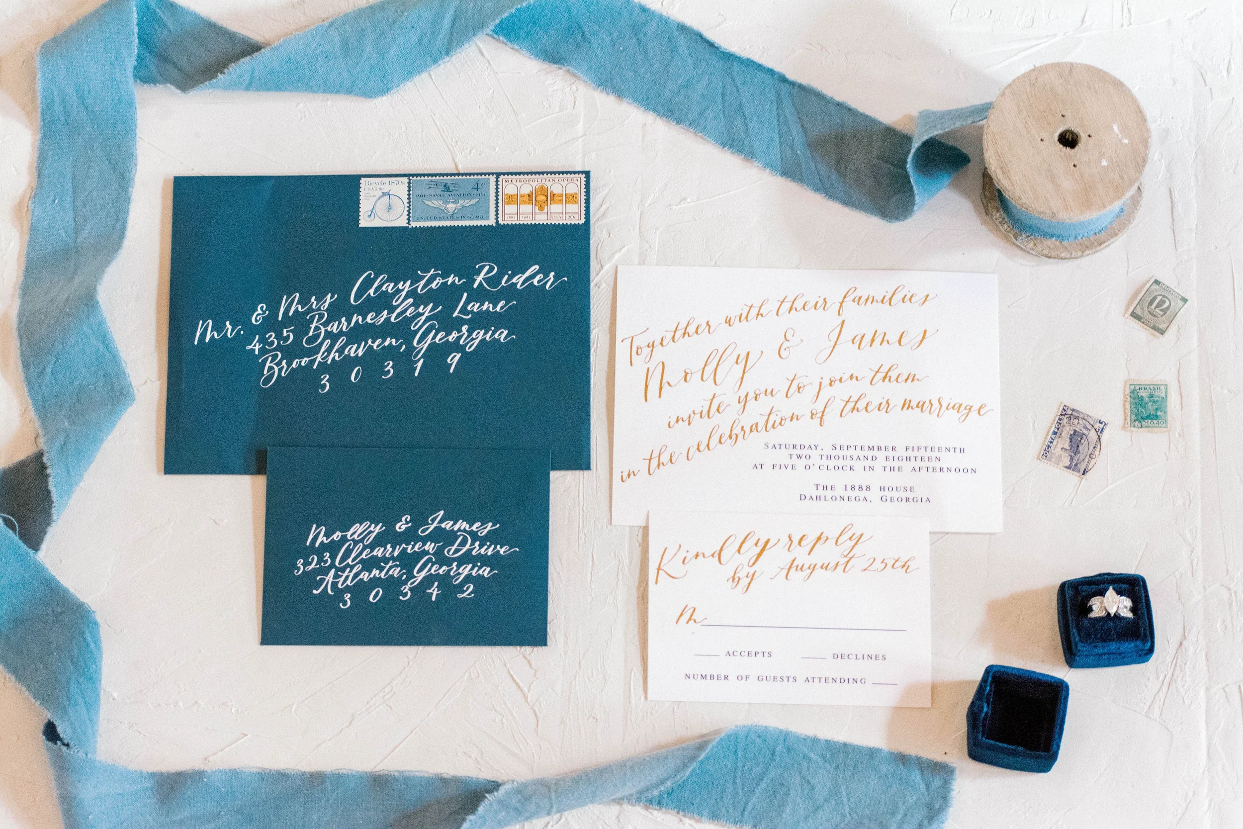 Make an impression before the wedding day with a striking blue and gold invitation suite. Hand-lettered in shimmering metallic gold ink on bright white cardstock, this fine-art stationery set features dark blue envelopes and sheer dusty blue silk rib