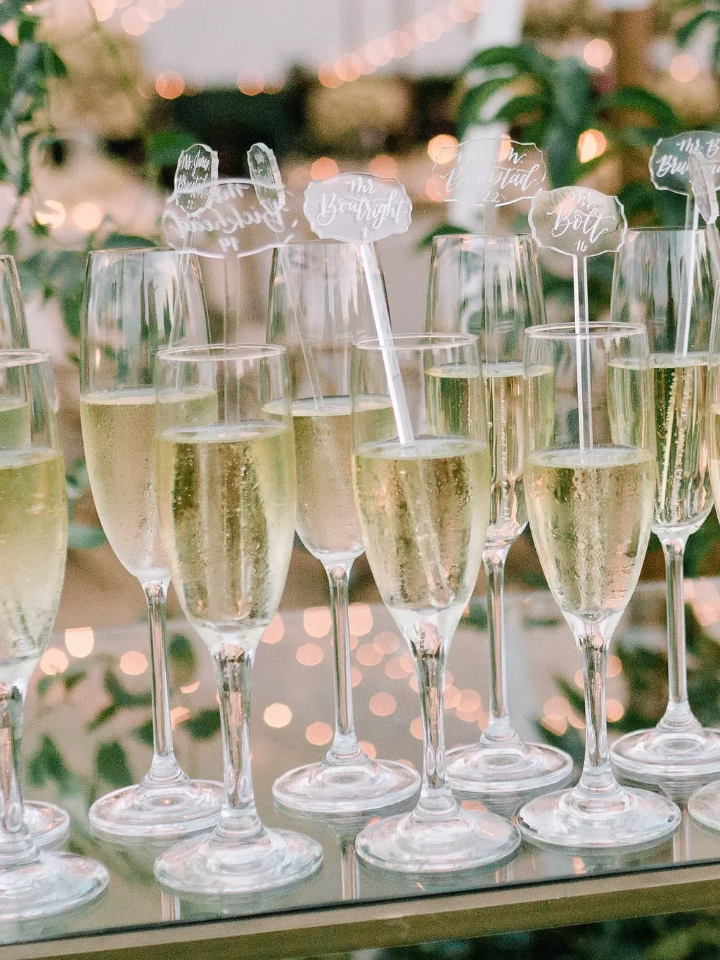 Close-up of champagne flutes filled with sparkling wine, each holding a small acrylic stir stick escort card with white calligraphy displaying guest names and table numbers, set on a glass shelf with soft bokeh string lights in the background.