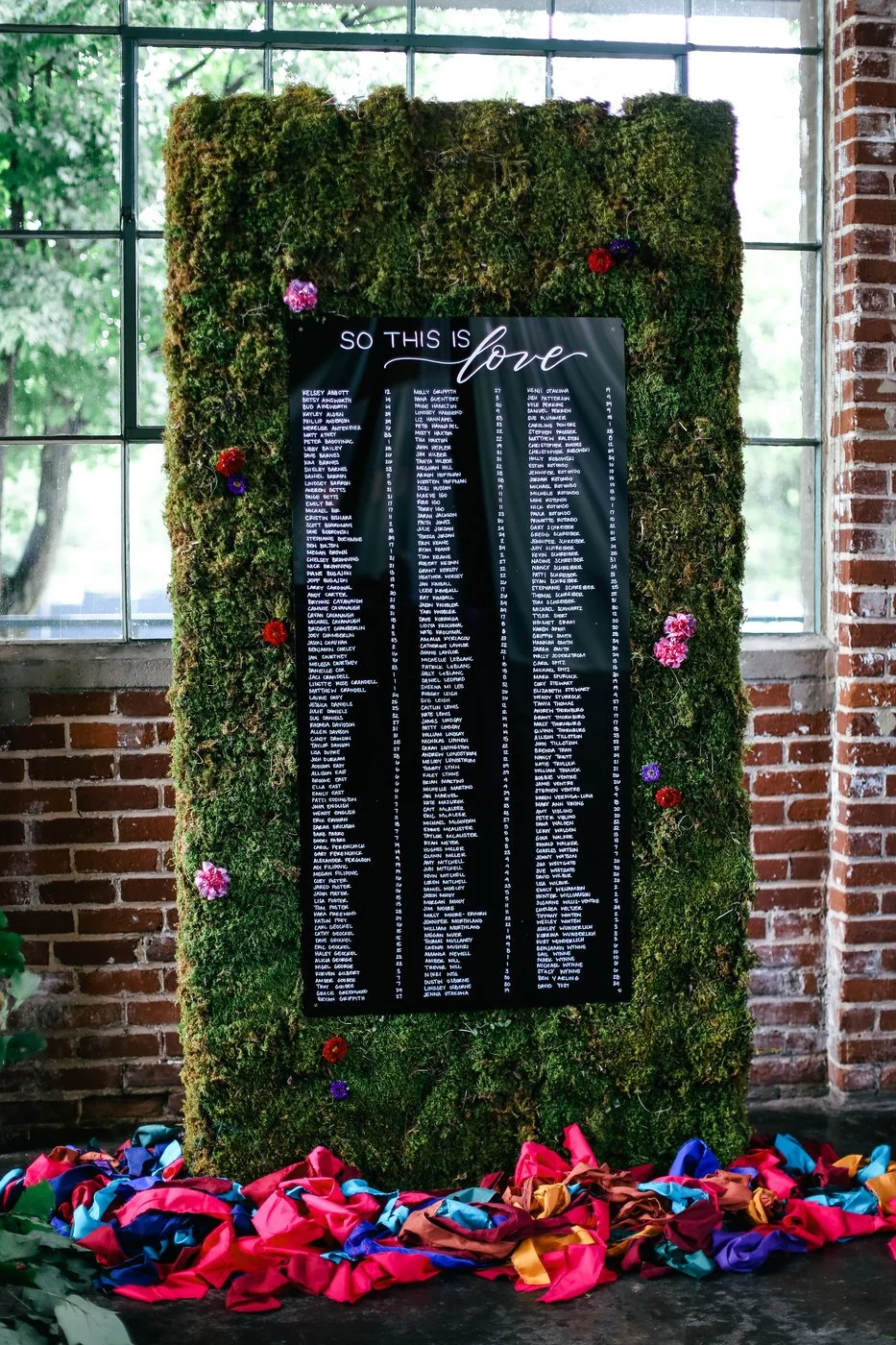 Black acrylic wedding seating chart with white calligraphy mounted on a moss backdrop with colorful flowers in an industrial venue.