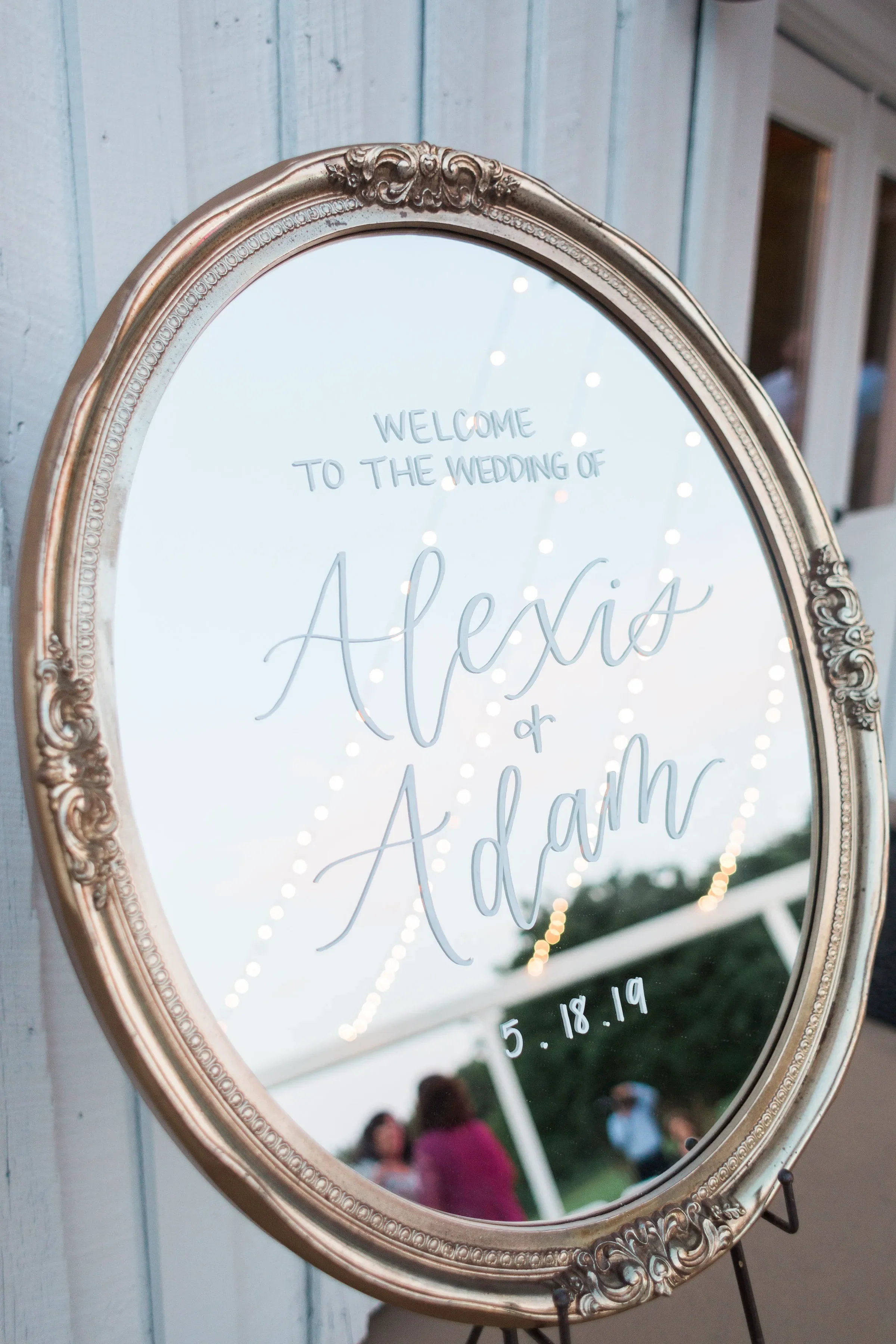 A large vintage gold oval framed mirror used as a wedding welcome sign with white script calligraphy displayed outdoors.