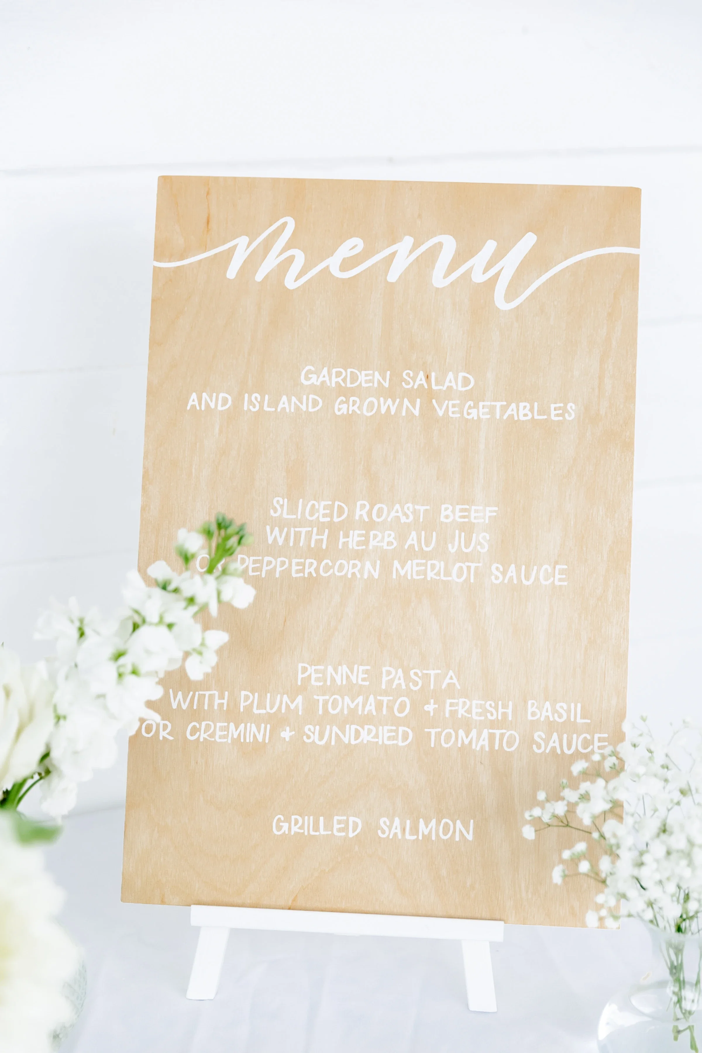 Replace individual paper menus with an eco-friendly wooden dinner menu sign. Showcasing clear white calligraphy on a light natural wood panel, this freestanding display gives guests a seamless, beautiful overview of your reception dining options.