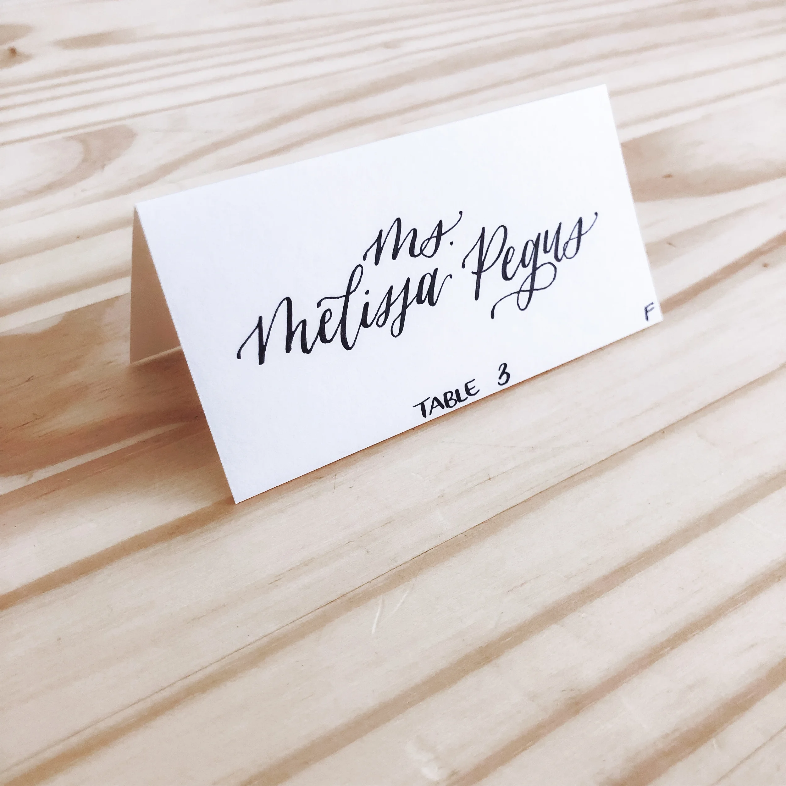 Embrace the timeless elegance of minimalist day-of stationery with a classic white folded place card. Showcasing clean, high-contrast black script calligraphy, this simple paper name card rests beautifully on natural wood or formal linen settings for