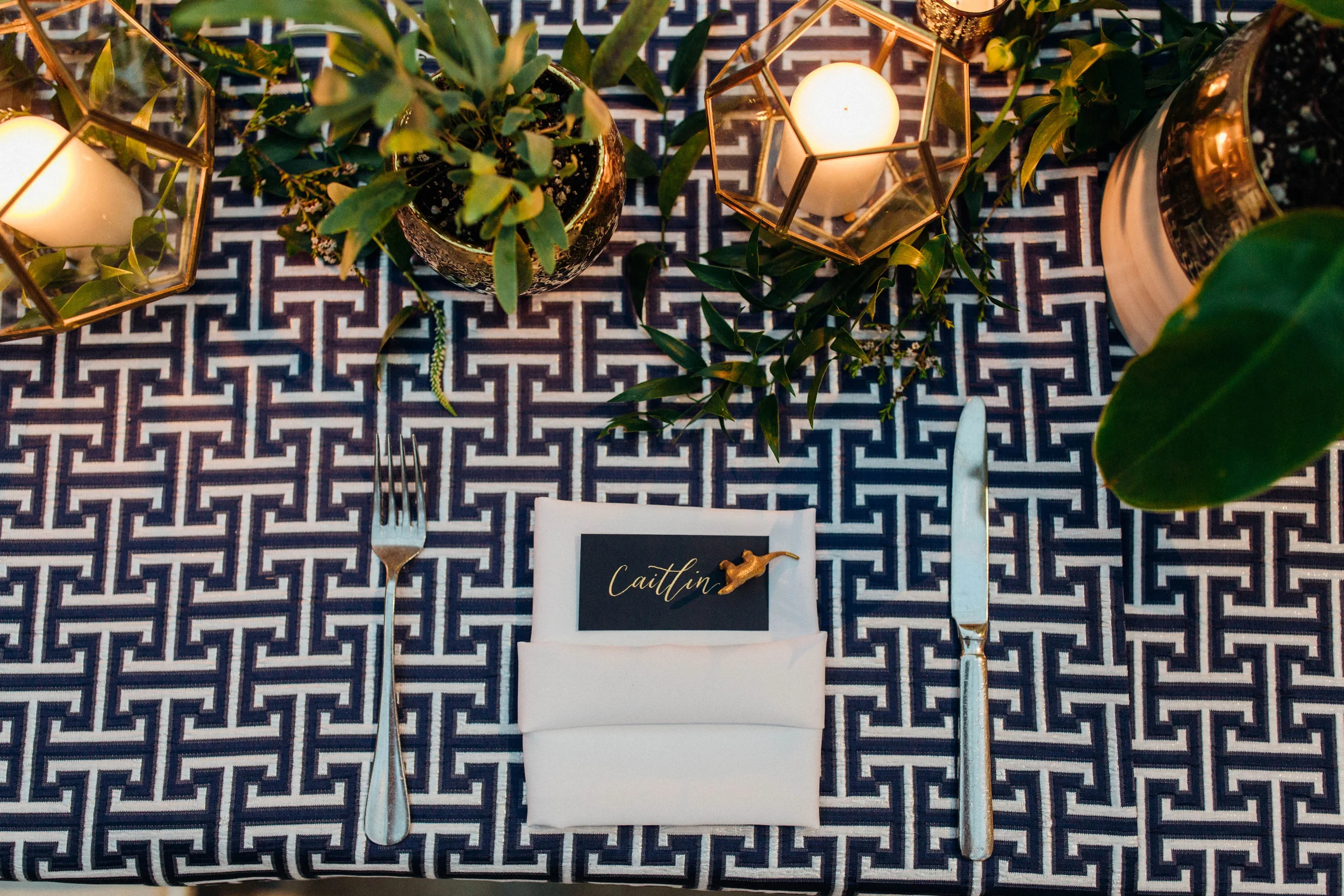 Navy blue and white geometric patterned tablecloth setting featuring a black paper place card with white cursive calligraphy and a small gold figure.