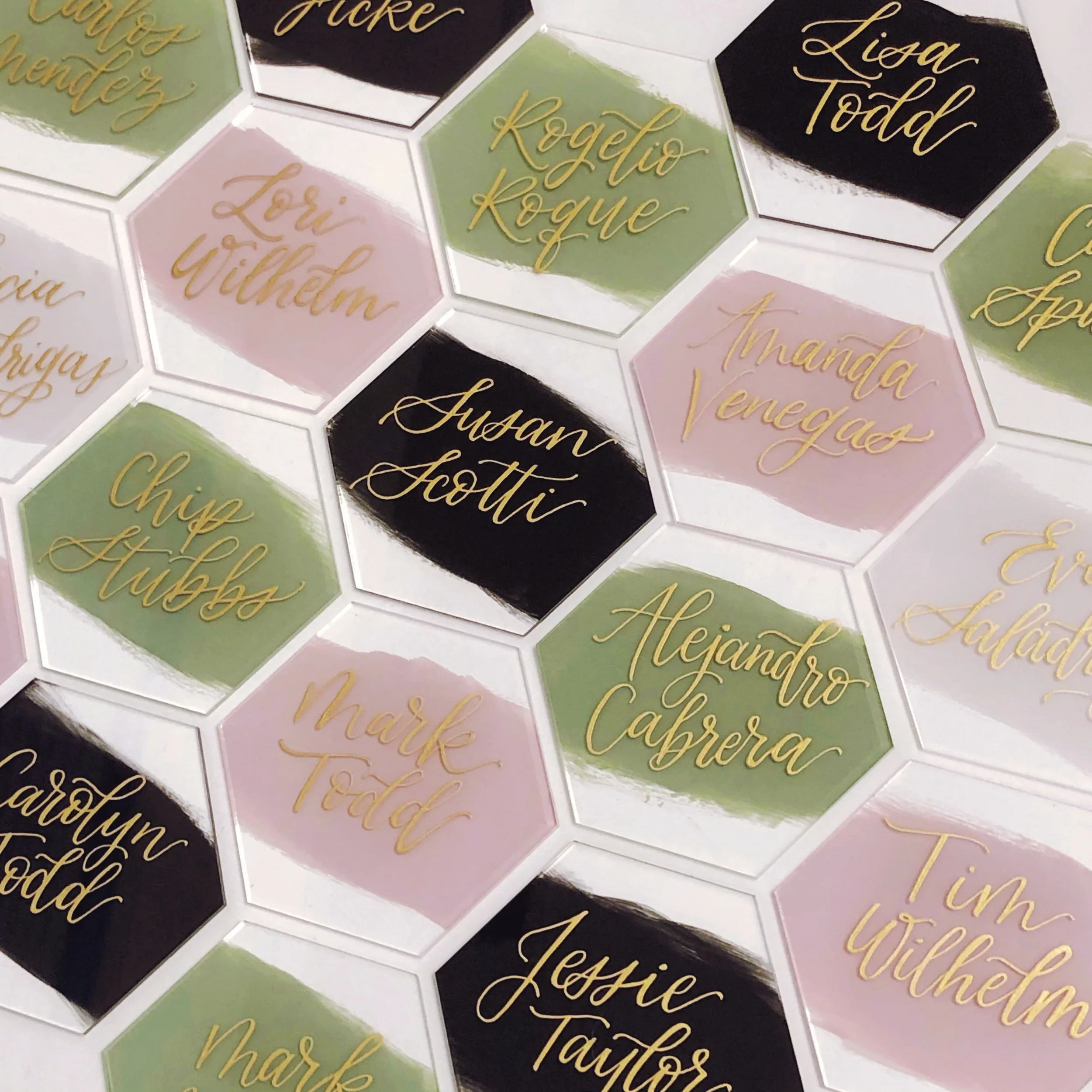 Make a bold geometric statement with a tiled array of painted hexagon place cards. Showcasing a sophisticated modern palette of sage green, black, and dusty blush pink, these custom-shaped escort tiles are finished with gleaming gold metallic calligr