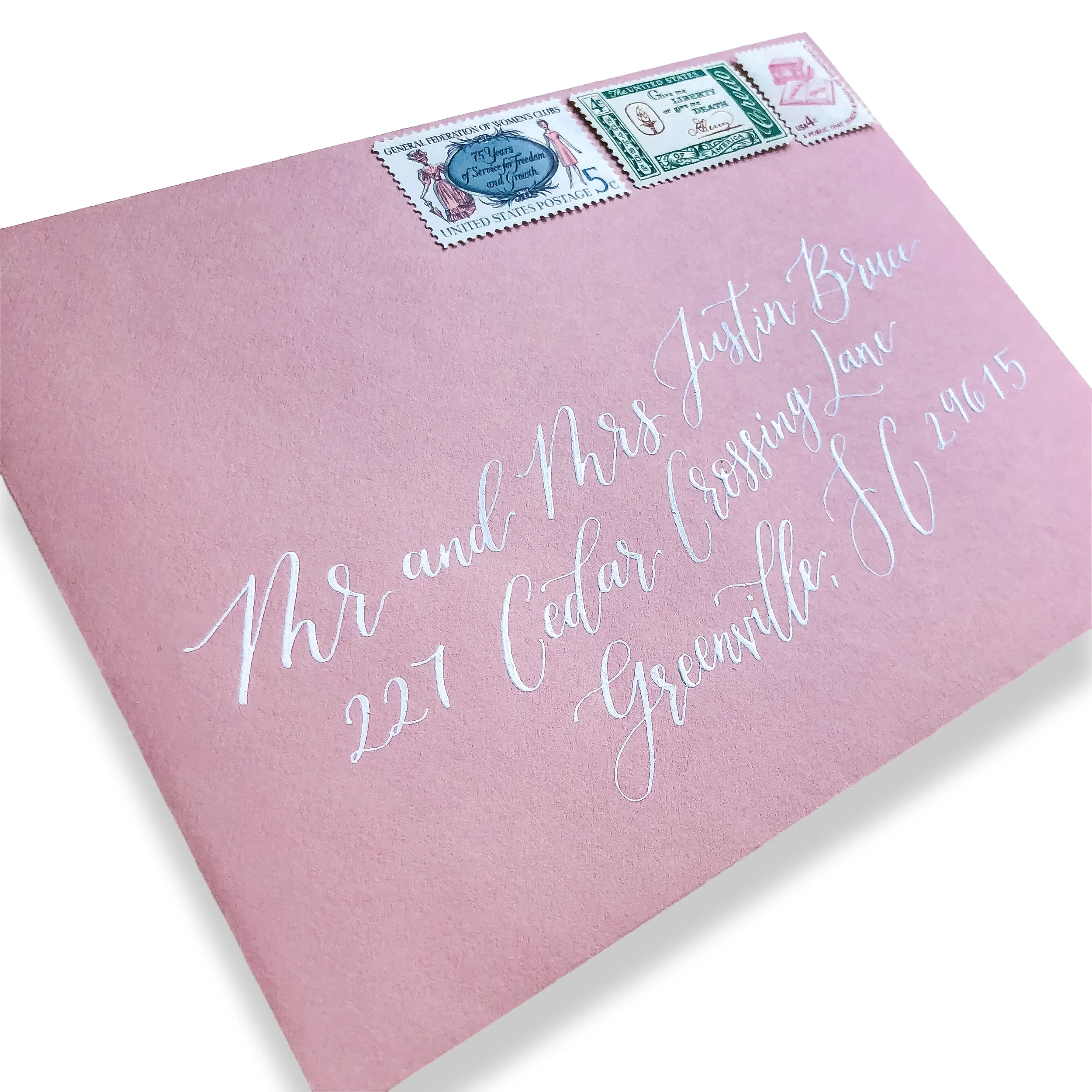 Dusty blush pink wedding envelope addressing in white calligraphy ink with an assortment of vintage postage stamps.