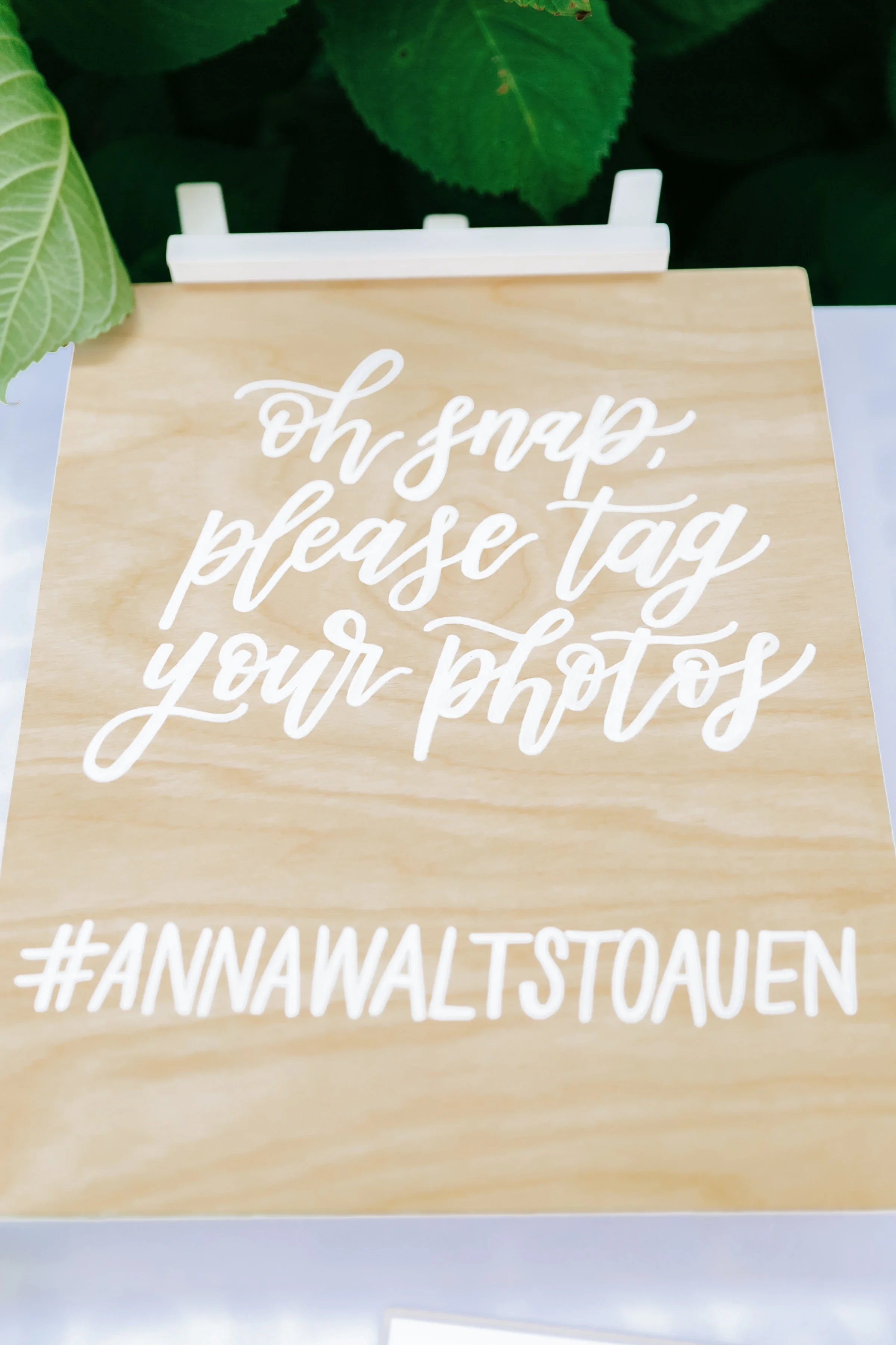 Hand-lettered wooden wedding hashtag sign displayed outdoors next to green leafy foliage.