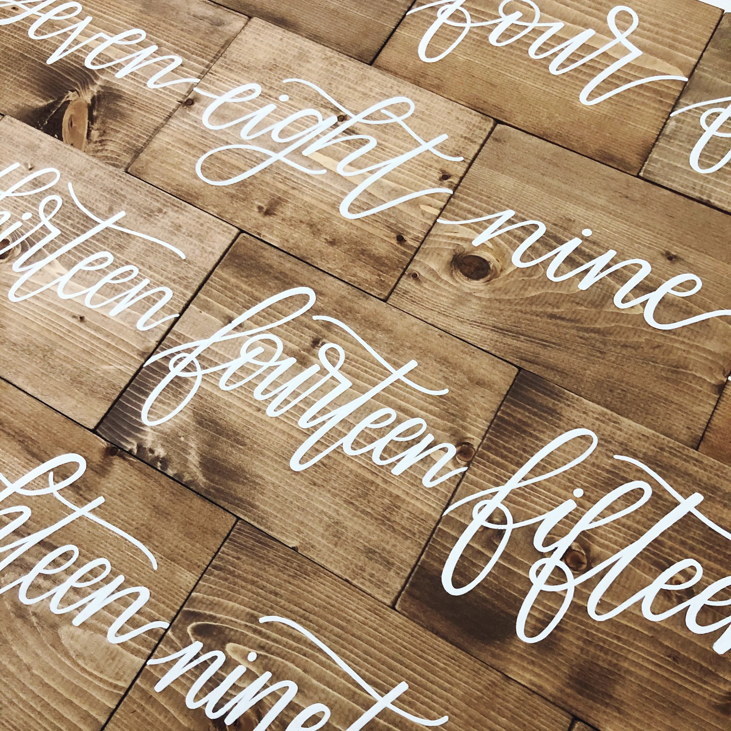 Add warm rustic charm to your reception with a freestanding stained wood block table number, lettered in bold white cursive script.