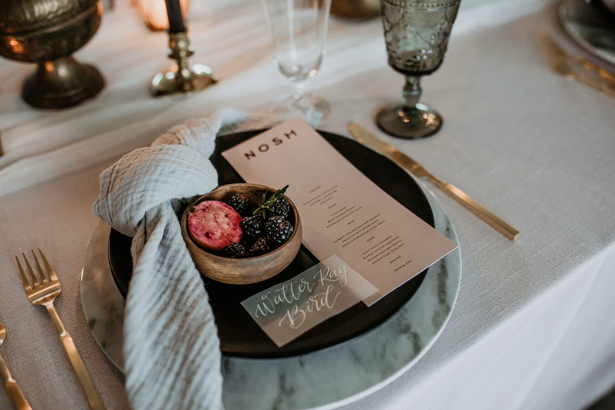 Fine art wedding table setting detailed with a vellum white ink calligraphy place card, blush dinner menu, sage linen napkin and a wooden bowl of sliced figs.
