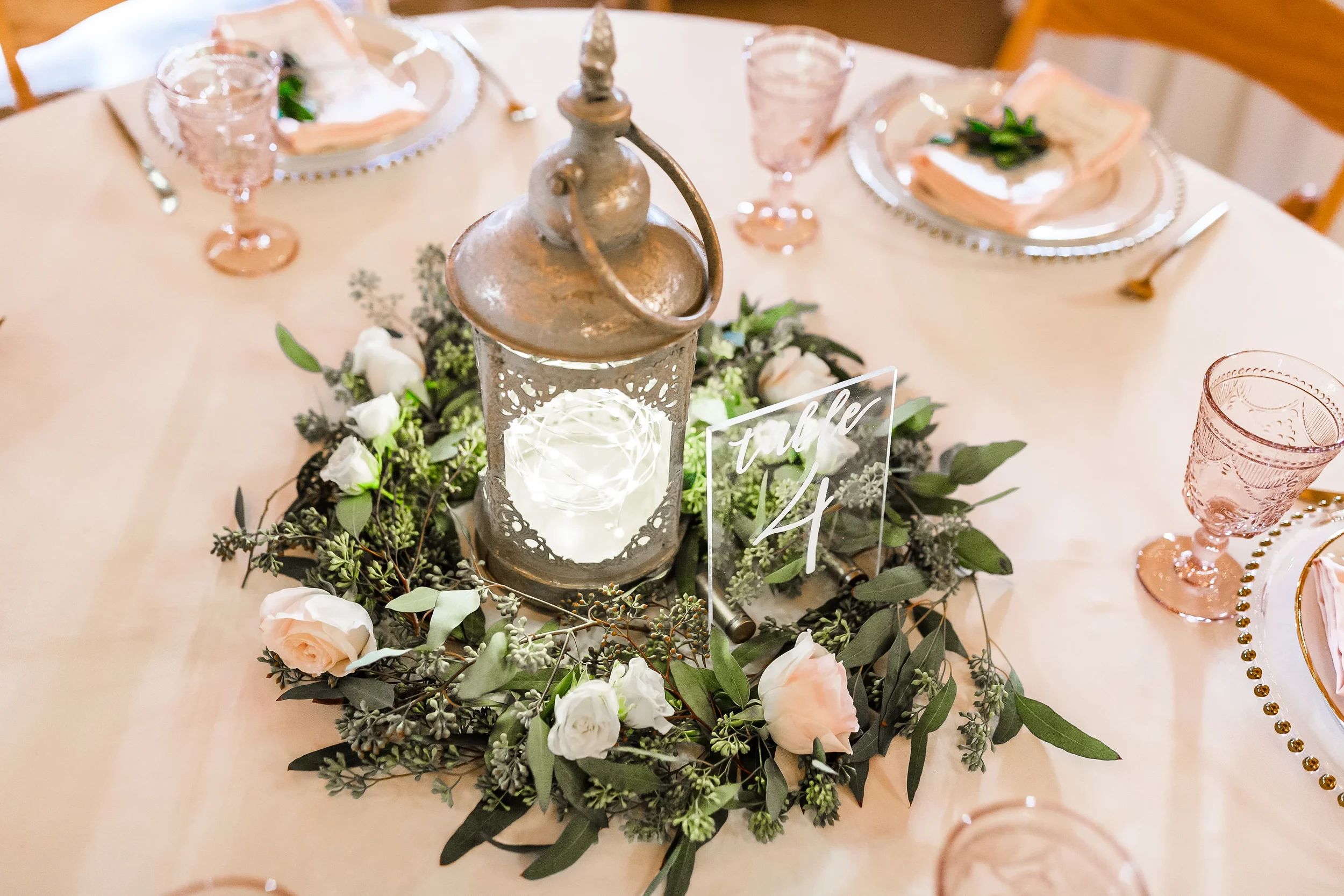 Modern clear acrylic table number 4 with white calligraphy script surrounded by a lantern and greenery centerpiece.