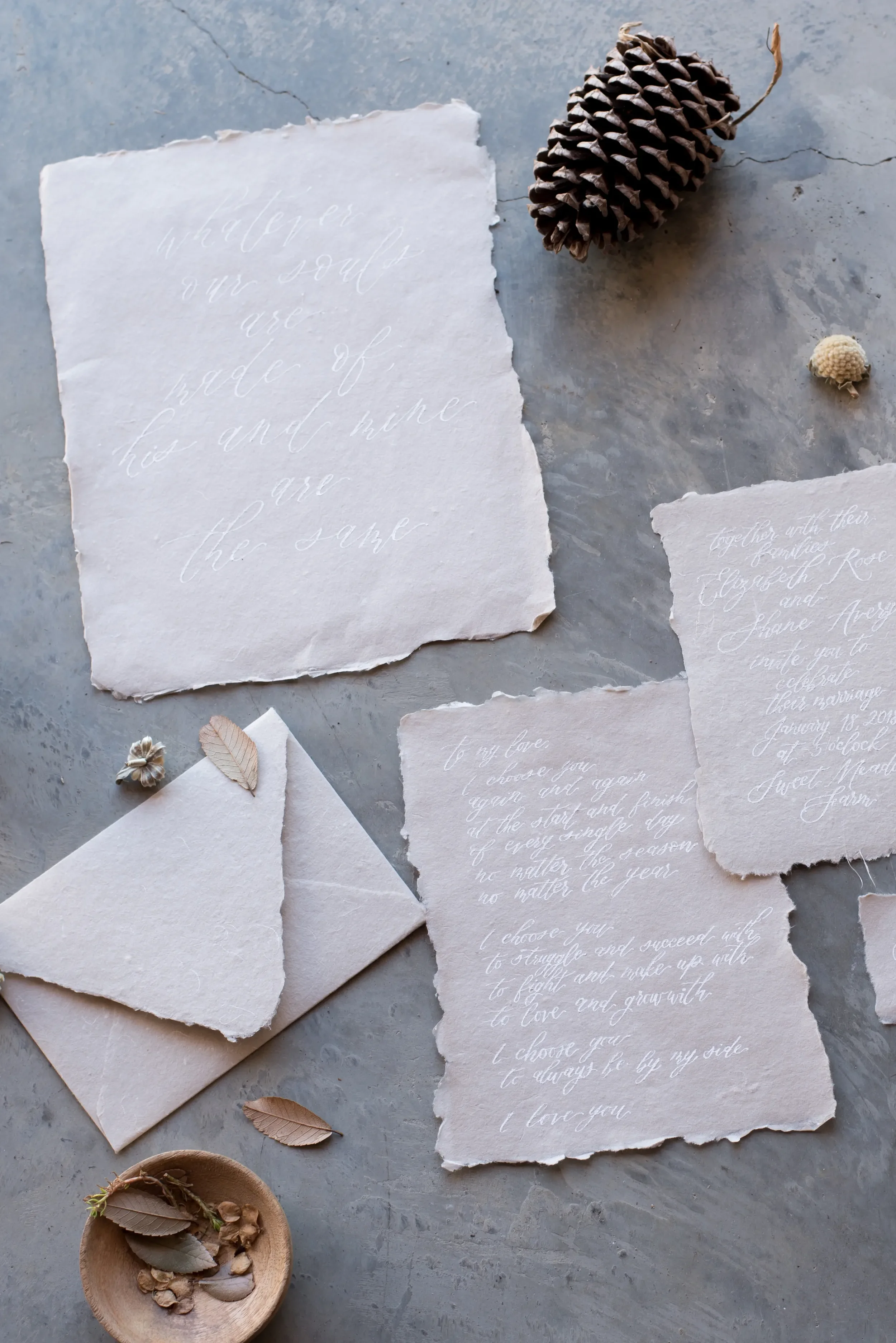 Fine art wedding stationery flat lay featuring white calligraphy on textured paper with natural foliage and pinecones.