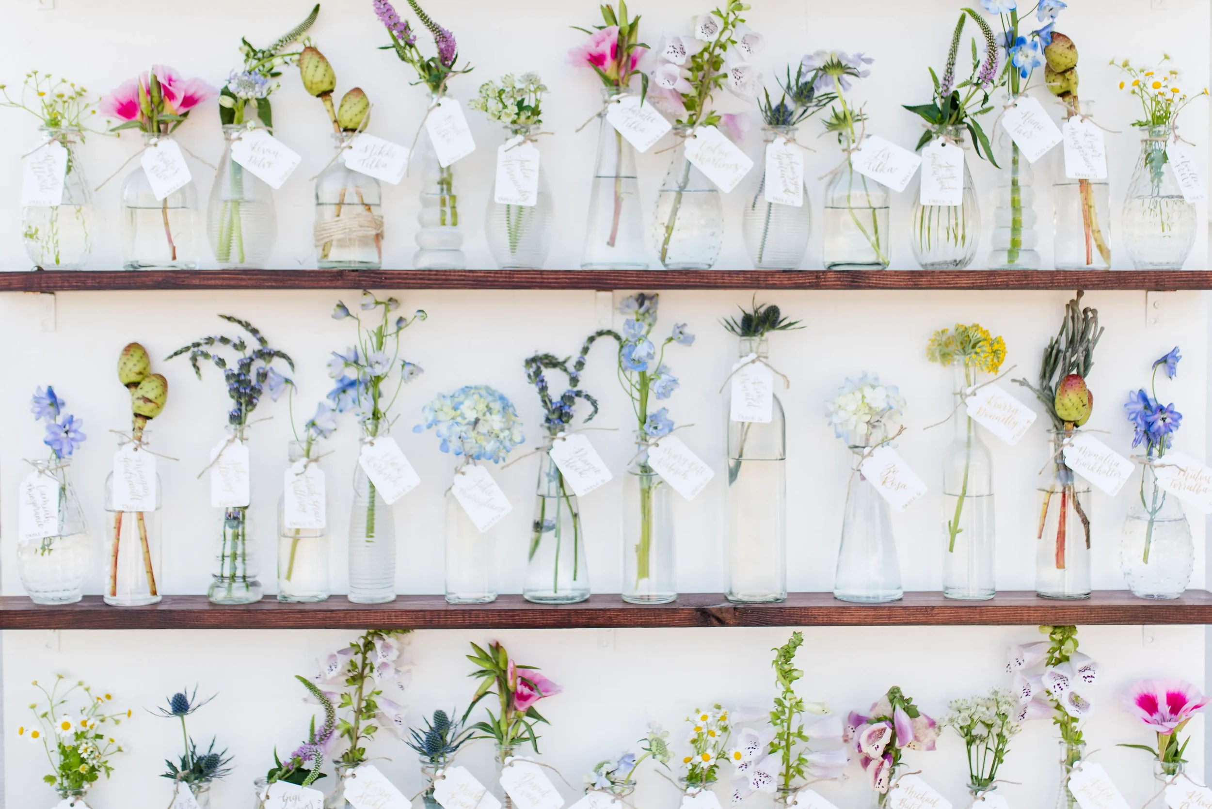 Delight your guests with a beautiful, multi-functional seating display that pairs delicate wildflower bud vases with custom calligraphy tags. This charming setup doubles as an interactive escort card wall and a sweet wedding favor for guests to take 