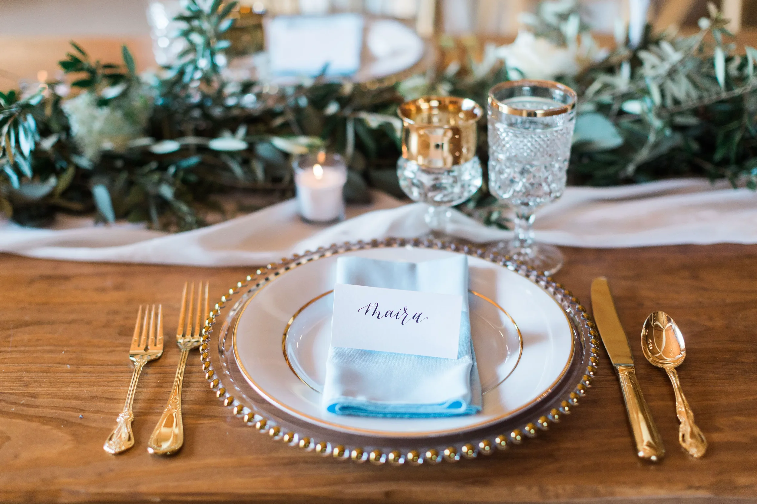 It is the fine details that truly welcome your guests. Give your reception tables a personalized, luxurious touch with custom handwritten script place cards. Beautifully paired here with soft blue textiles, elegant gold accents, crystal glassware, an