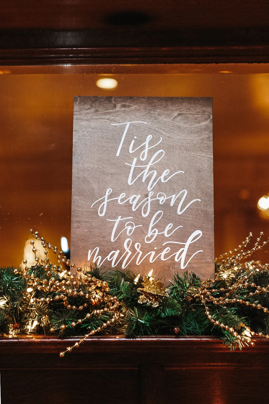 Dark wood sign hand-lettered in white calligraphy reading "Tis the season to be married" set on a decorated mantel.