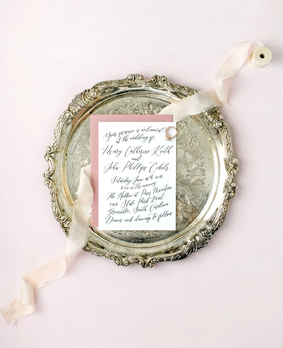 Hand-calligraphed wedding invitation card on a blush envelope, styled on an antique silver tray with flowing blush silk ribbon and a diamond ring.
