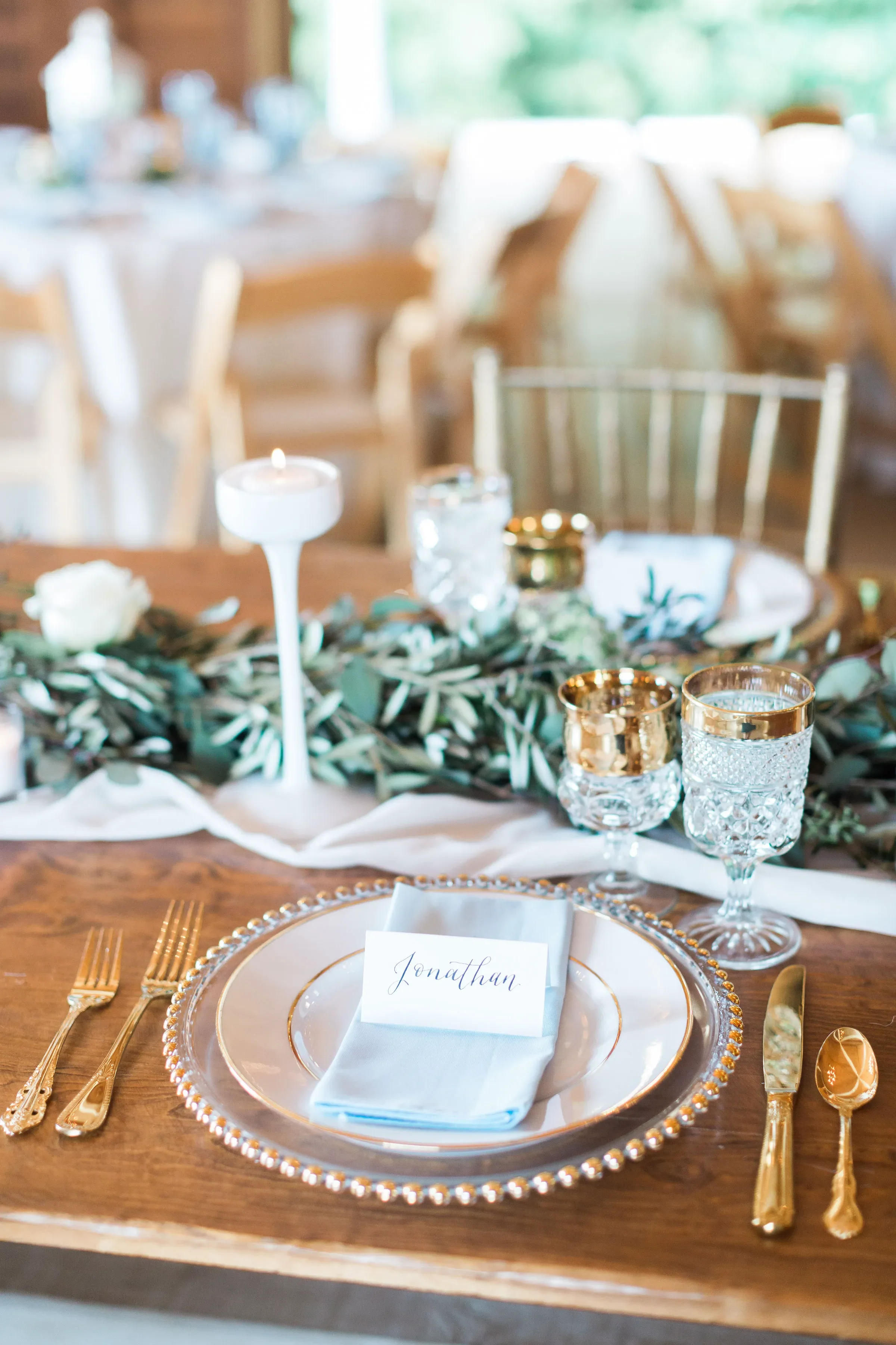 Create a luxurious dining experience with elegant handwritten calligraphy place cards. This custom paper escort card features delicate script lettering, beautifully styled on a soft light blue napkin with gold-rimmed dinnerware and a clear glass bead