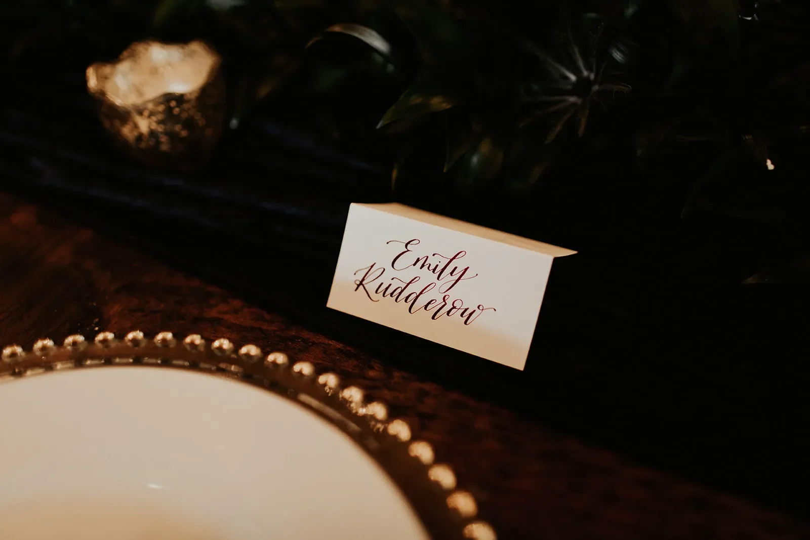 Calligraphy tented place cards with burgundy lettering in front of a gold rimmed charger for a moody autumn wedding.