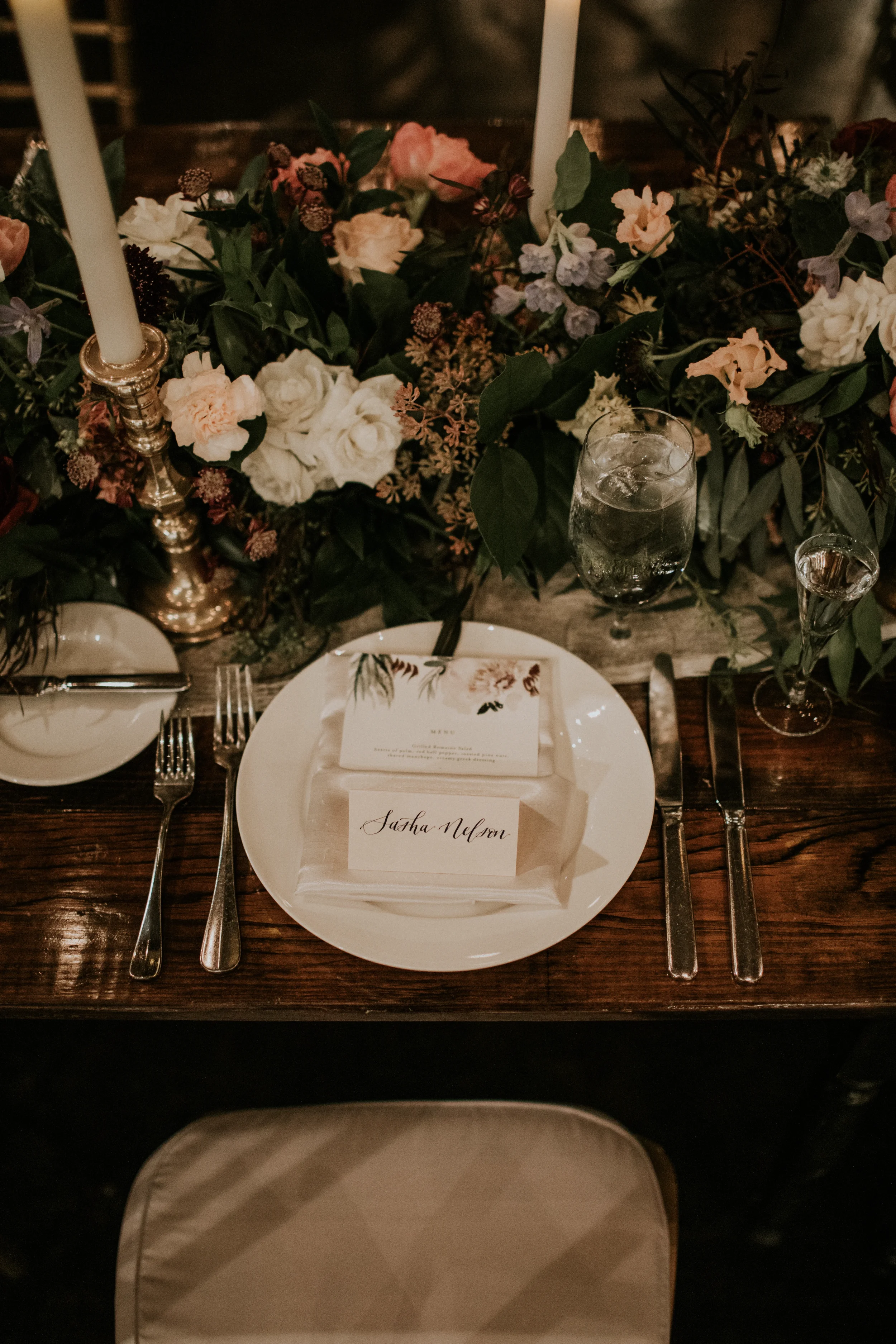 Top-down view of a rustic wooden wedding reception table setting featuring a handwritten calligraphy place card reading "Sasha Nelson" resting on a white napkin and menu card, surrounded by silver silverware, fine glassware, and a lush garden floral 